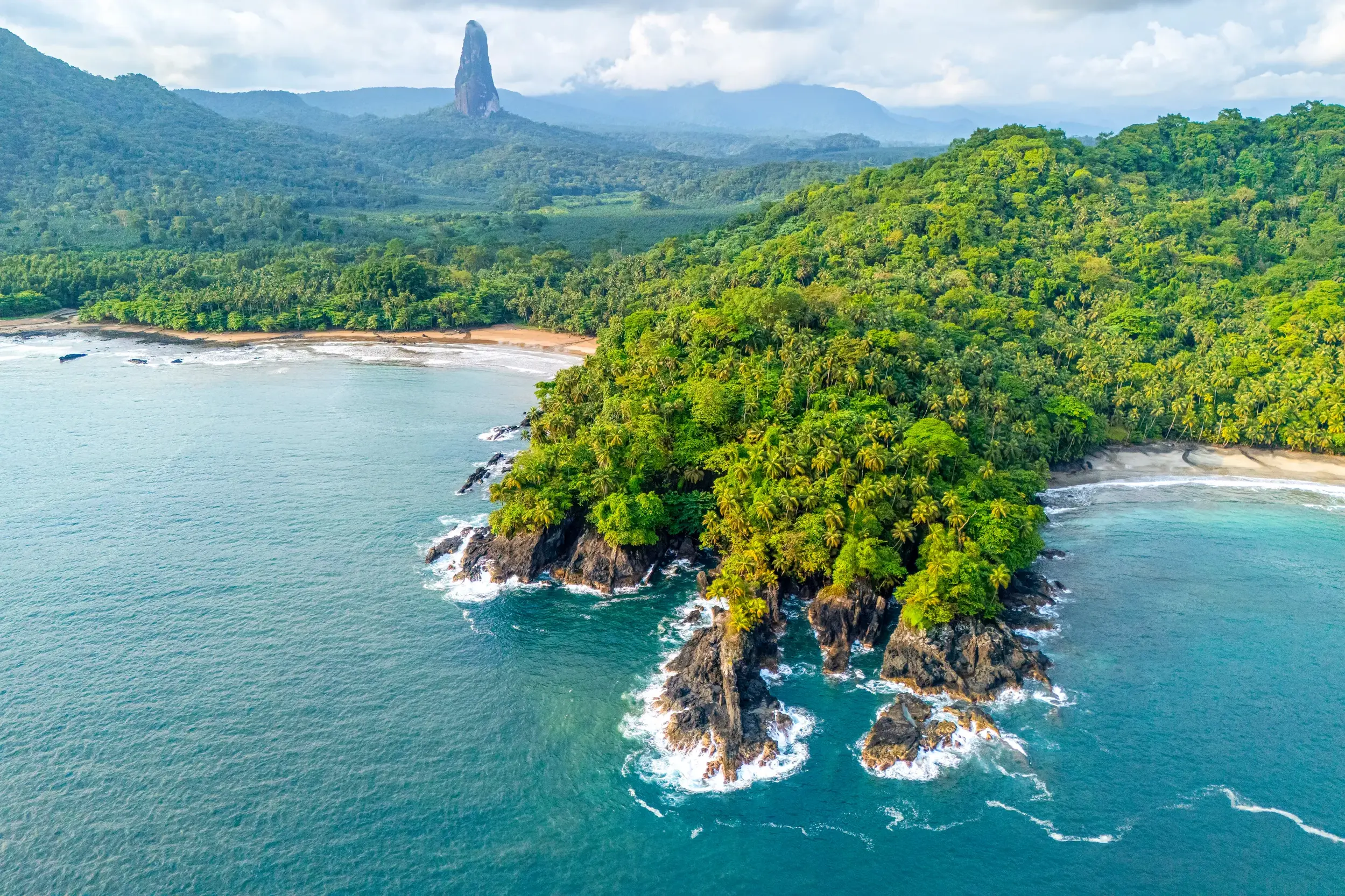 Luftaufnahme der wilden, bewaldeten Küste von São Tomé mit Blick auf den markanten Vulkanfelsen Pico Cão Grande am Horizont.