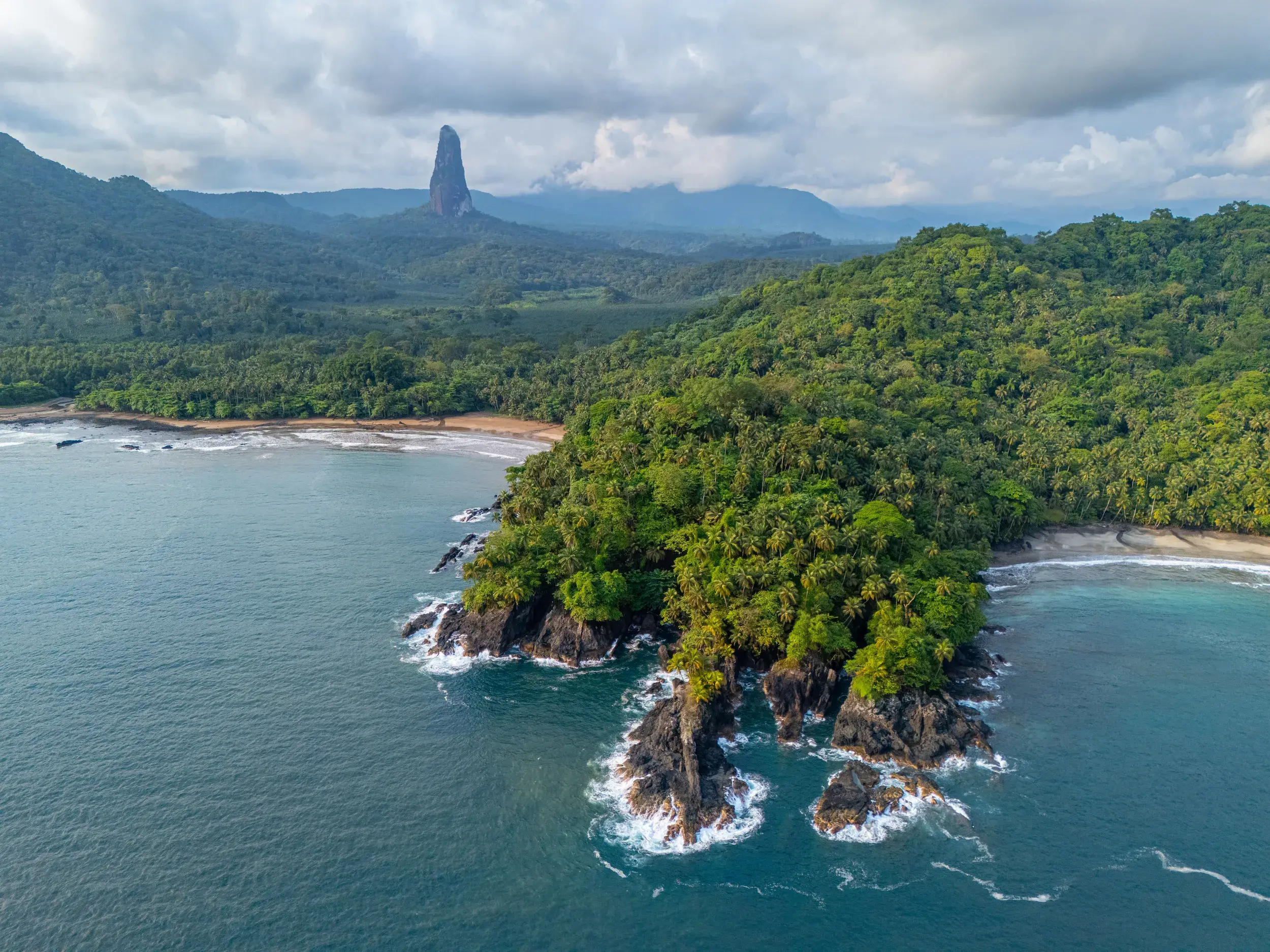 Luftaufnahme der wilden, bewaldeten Küste von São Tomé mit Blick auf den markanten Vulkanfelsen Pico Cão Grande am Horizont.