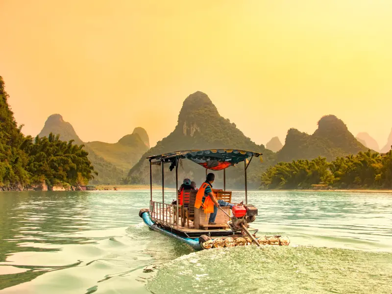Zwei Reisende auf einer Bootsfahrt auf dem Li-Fluss bei Yangshuo, vorbei an den grünen Zuckerhutbergen im Abendlicht.