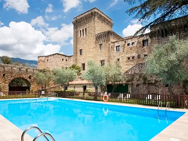 Leuchtend blauer Swimmingpool vor der historischen Steinfassade des Parador de Jarandilla de la Vera unter wolkigem Himmel.