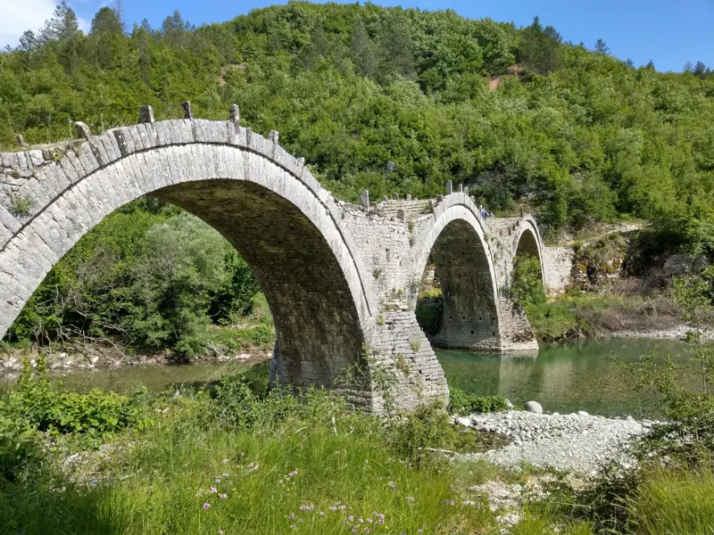Wanderer überqueren eine historische Steinbogenbrücke mit mehreren Bögen über dem Voidomatis-Fluss in der grünen Zagori-Region.