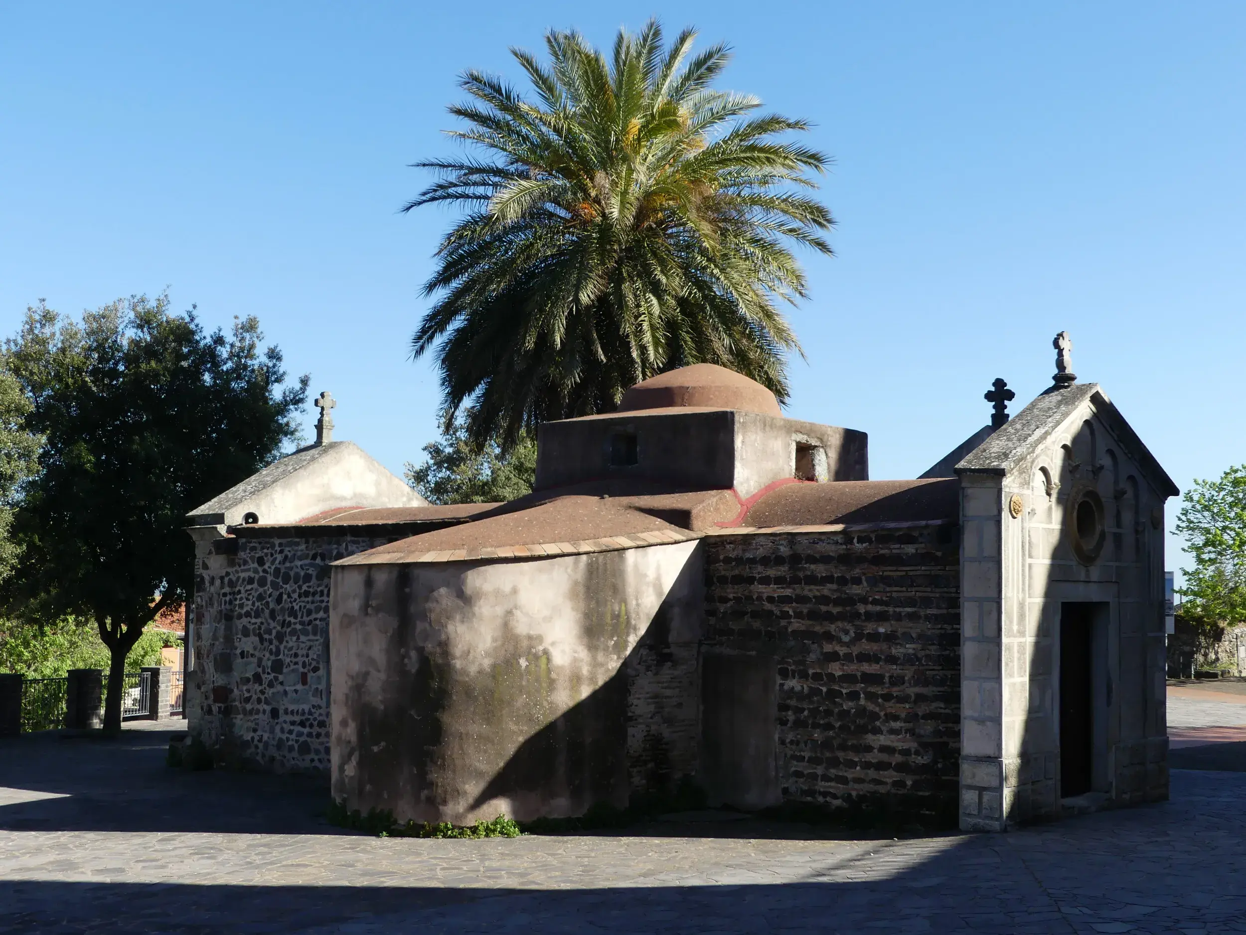 Rustikale Steinkirche in Bonarcado auf Sardinien mit Kuppeldach und Kreuzen an einem sonnigen Tag unter einer Palme.