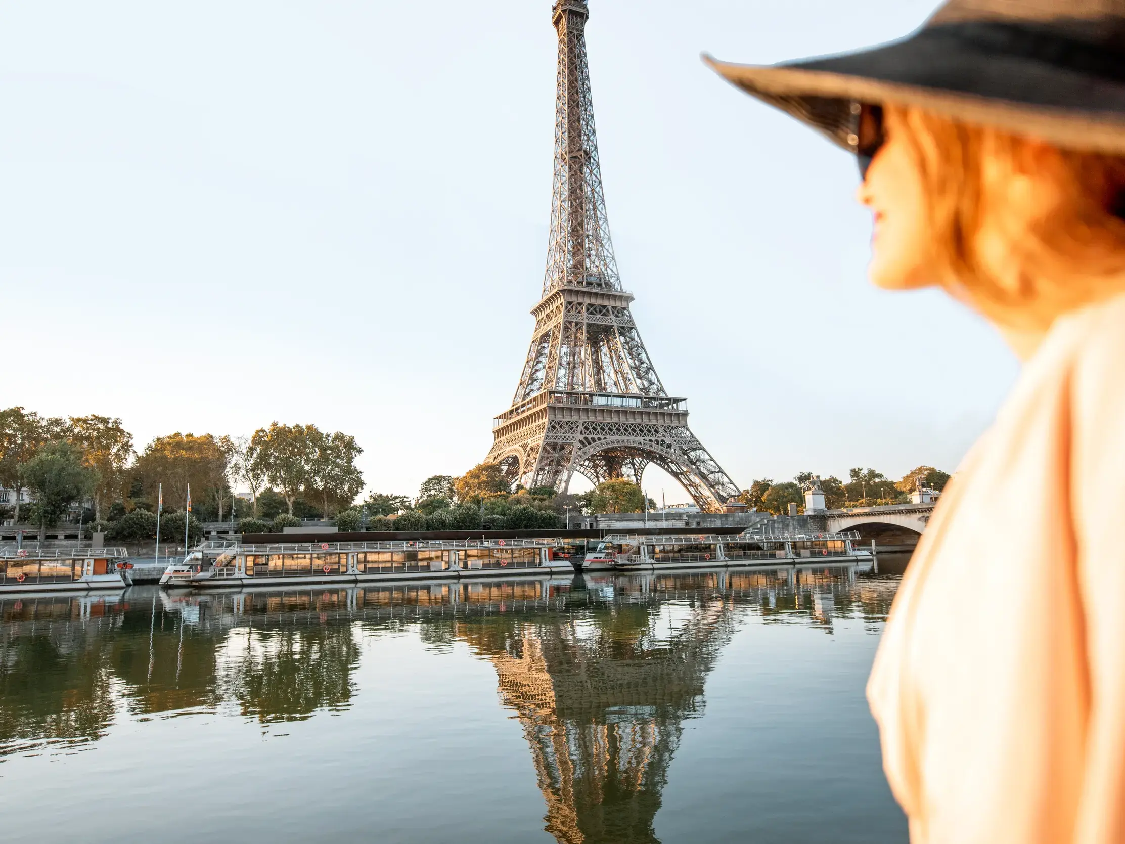 Eine Frau mit Hut blickt auf den Eiffelturm in Paris, der sich im Wasser der Seine neben Ausflugsbooten spiegelt.