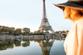 Eine Frau mit Hut blickt auf den Eiffelturm in Paris, der sich im Wasser der Seine neben Ausflugsbooten spiegelt.