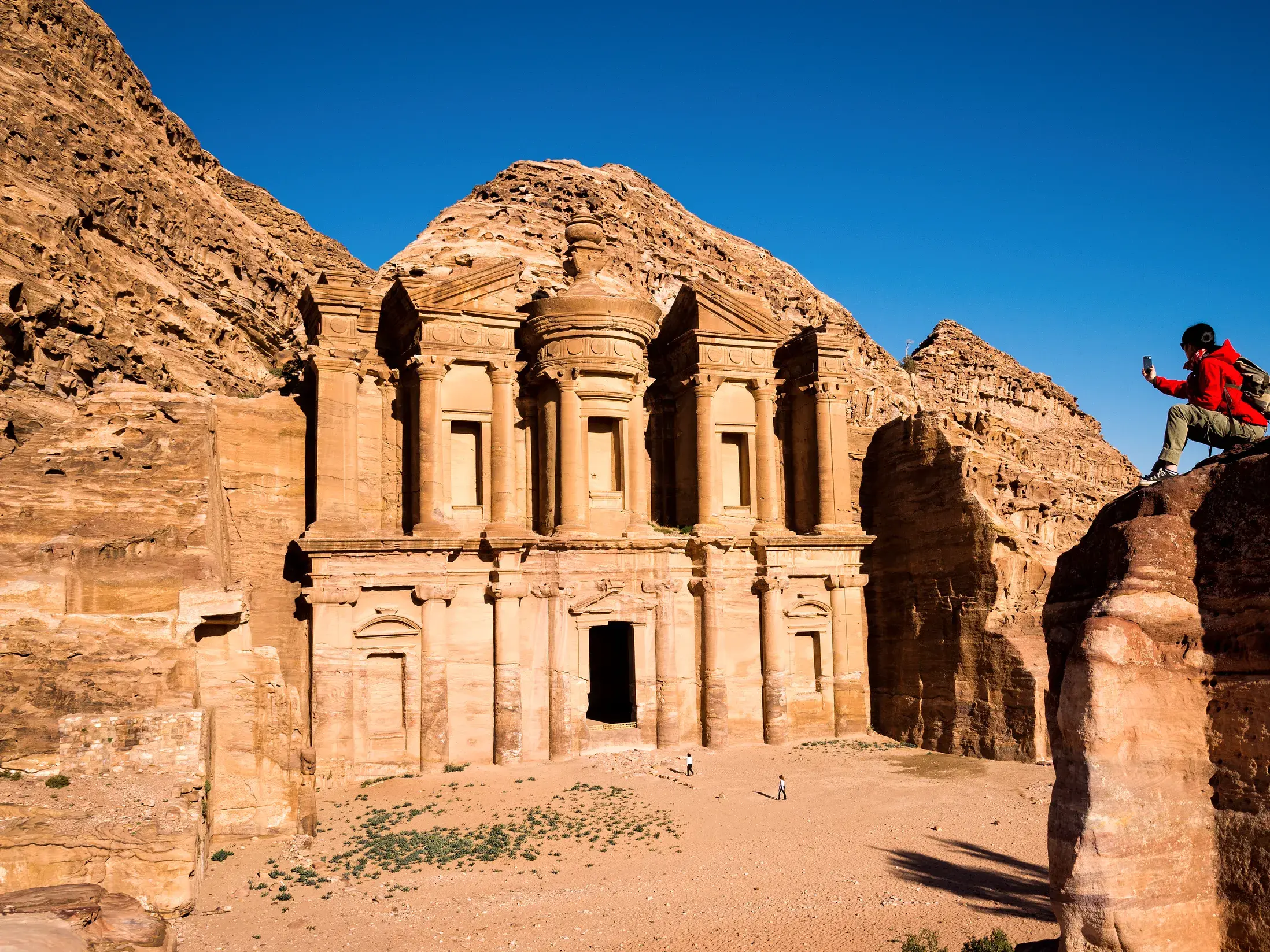 Ein Reisender in roter Jacke fotografiert von einem Felsen aus das monumentale Kloster Ad-Deir in der Felsenstadt Petra.