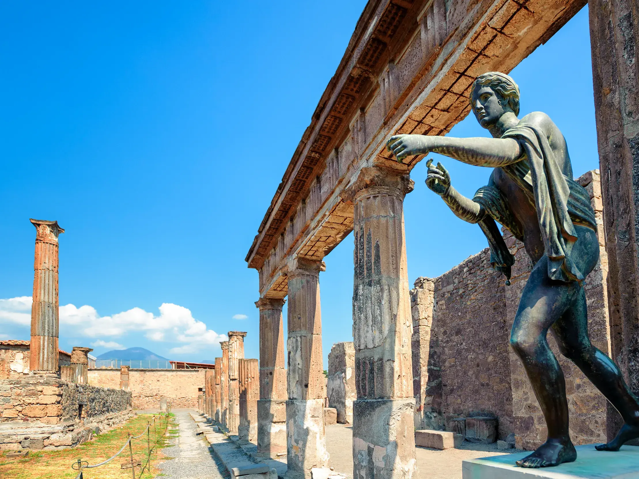 Bronzestatue neben einer Säulenreihe in den antiken Ruinen von Pompeji mit Blick auf den Vesuv an einem sonnigen Tag.