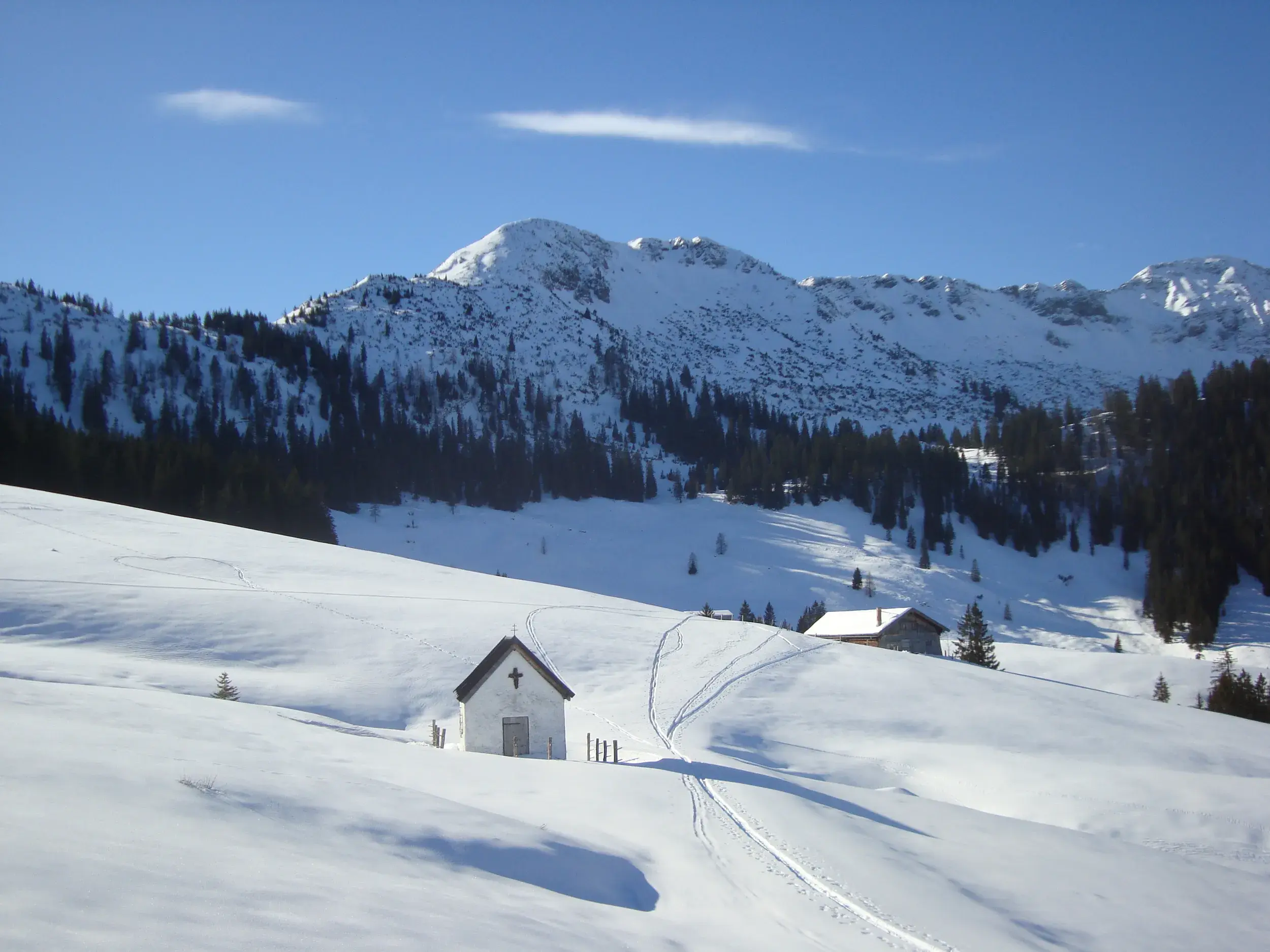 Eine Kapelle im tiefen Schnee im Risstal. Spuren einer Schneeschuhwanderung führen zum sonnigen Bergpanorama des Karwendel.