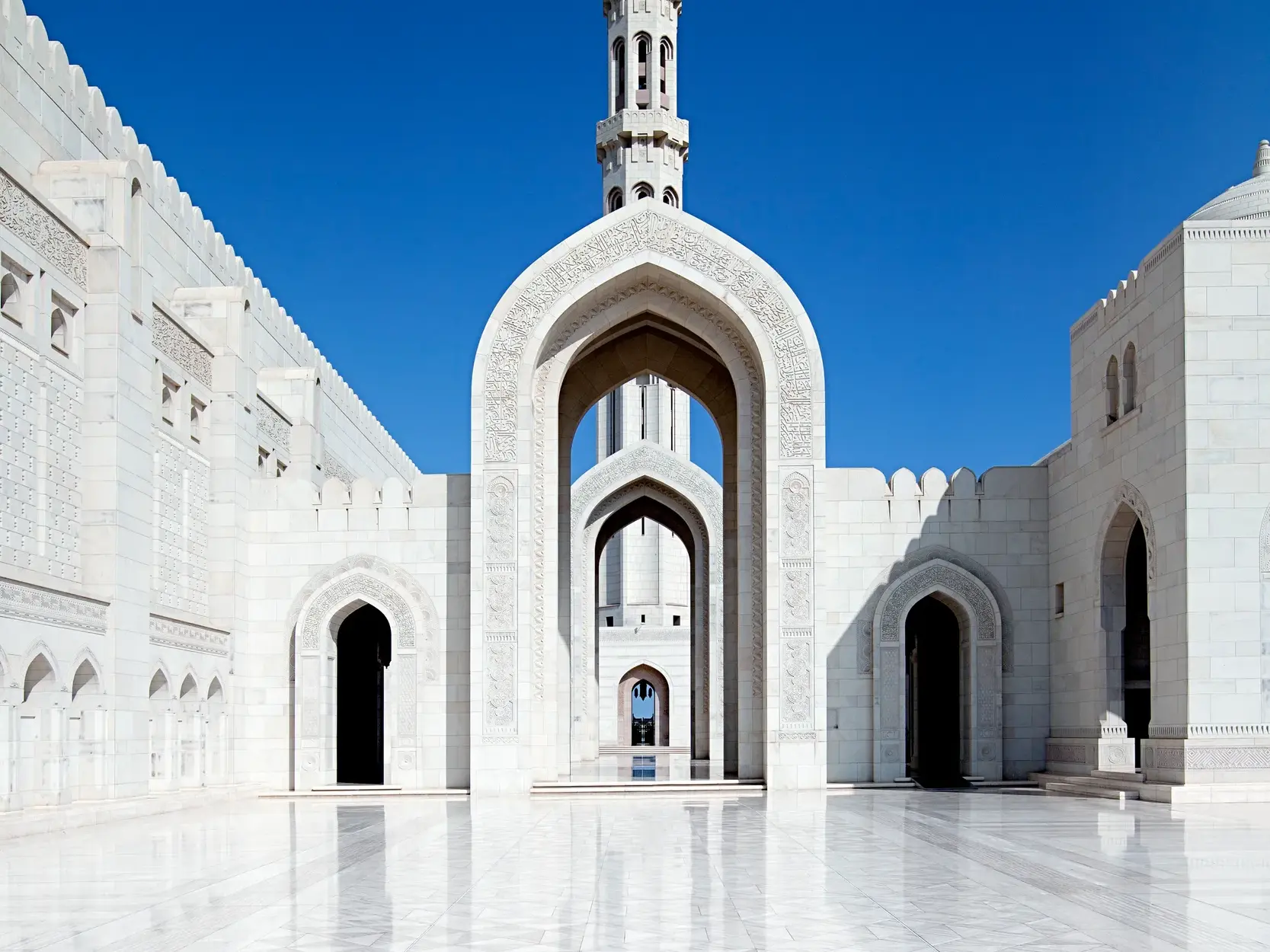 Der polierte Marmor-Innenhof der Sultan-Qaboos-Moschee in Maskat mit kunstvoll verzierten, weißen Arkaden unter blauem Himmel.
