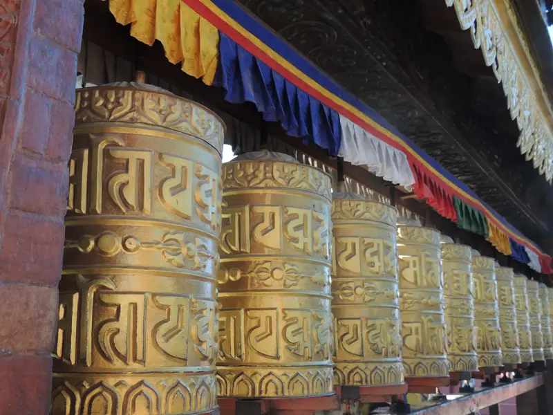 Aufgereihte goldene Gebetsmühlen mit Gravuren in einem Kloster in Nepal, darüber hängen bunte Gebetsfahnen.