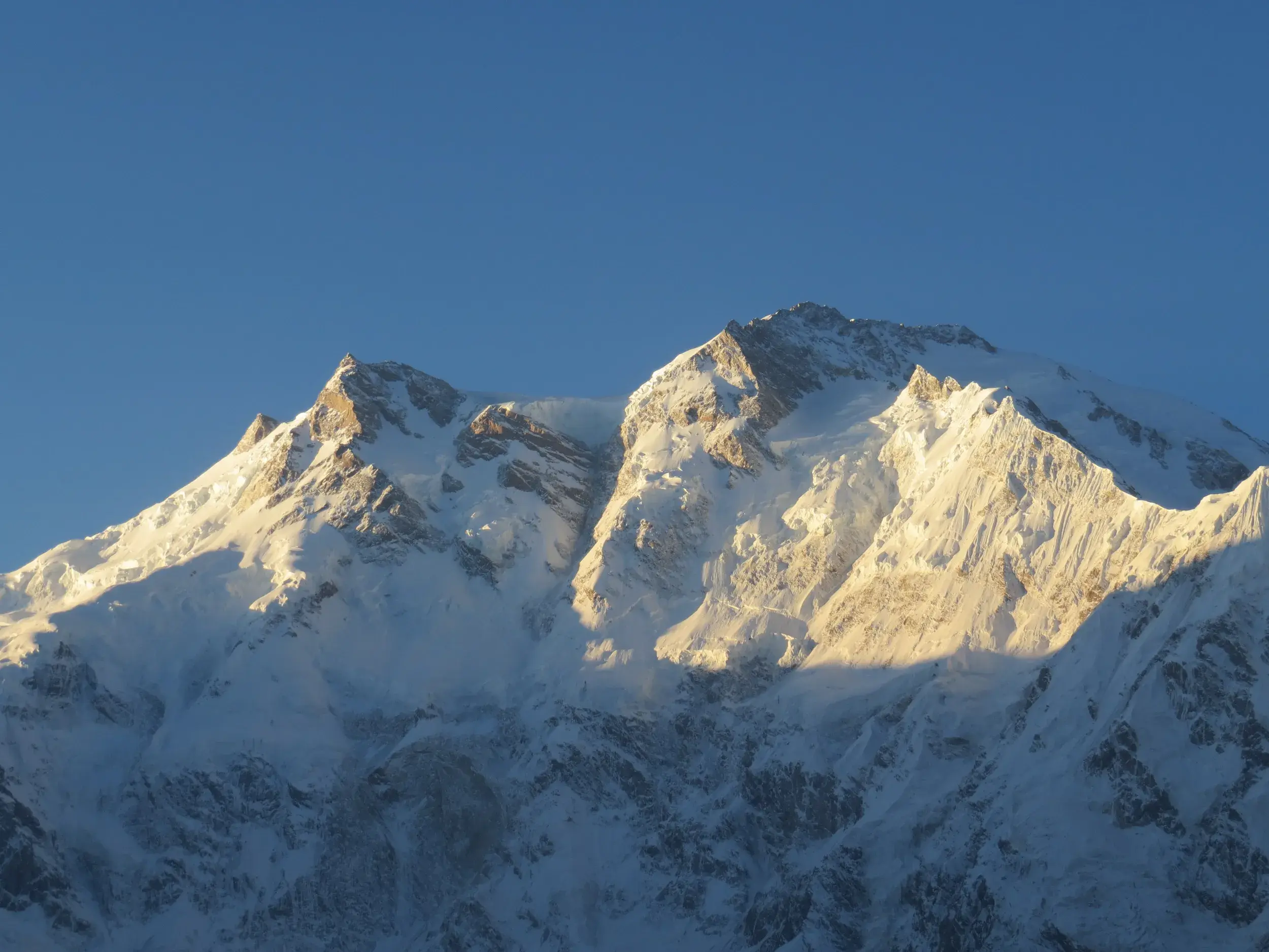 Die schneebedeckten, felsigen Gipfel des Nanga Parbat in Pakistan leuchten im warmen Morgenlicht vor einem klaren, blauen Himmel.