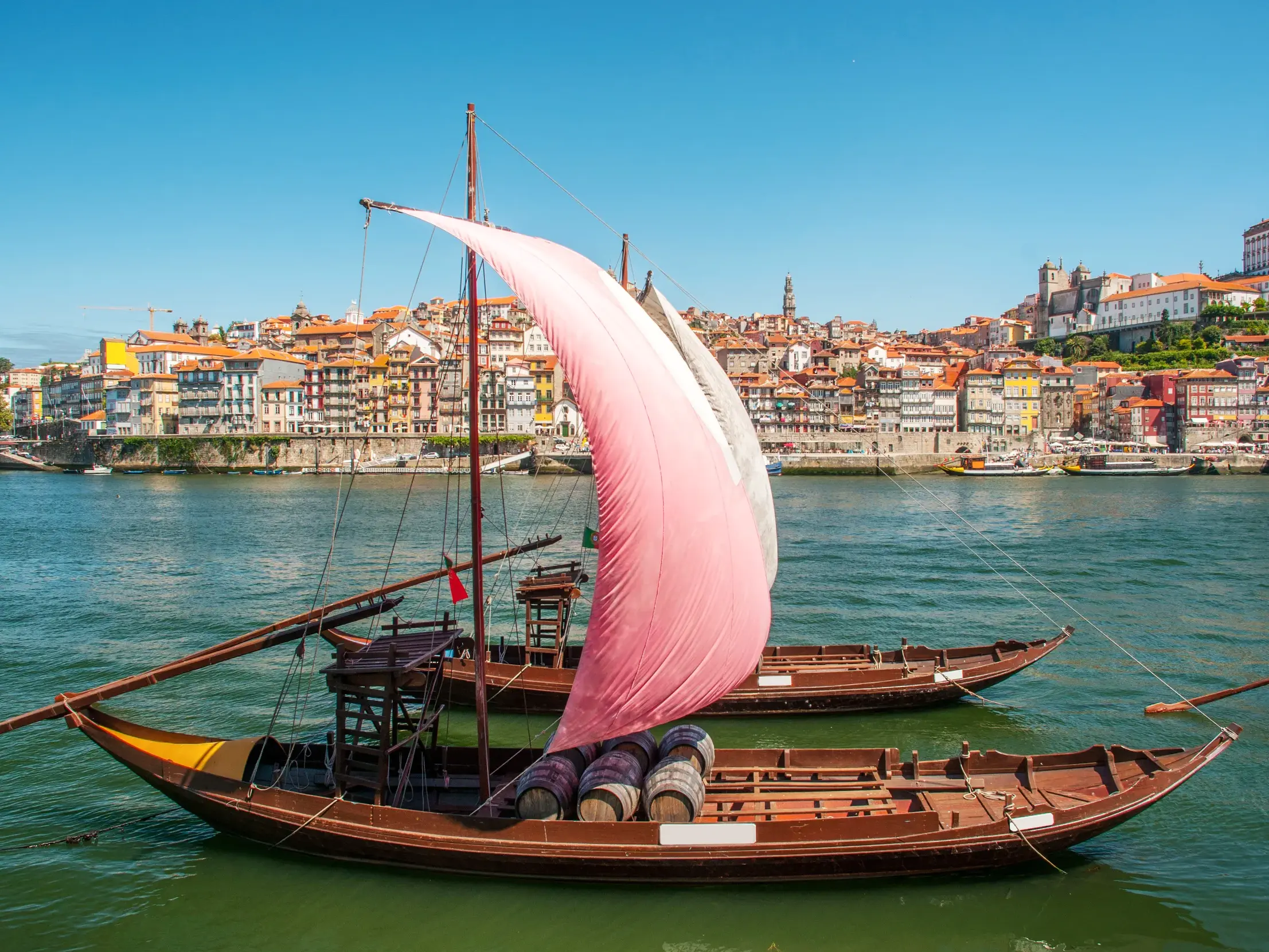 Zwei traditionelle Rabelo-Boote mit Weinfässern auf dem Fluss Douro vor der farbenfrohen Altstadt von Porto bei Sonnenschein.