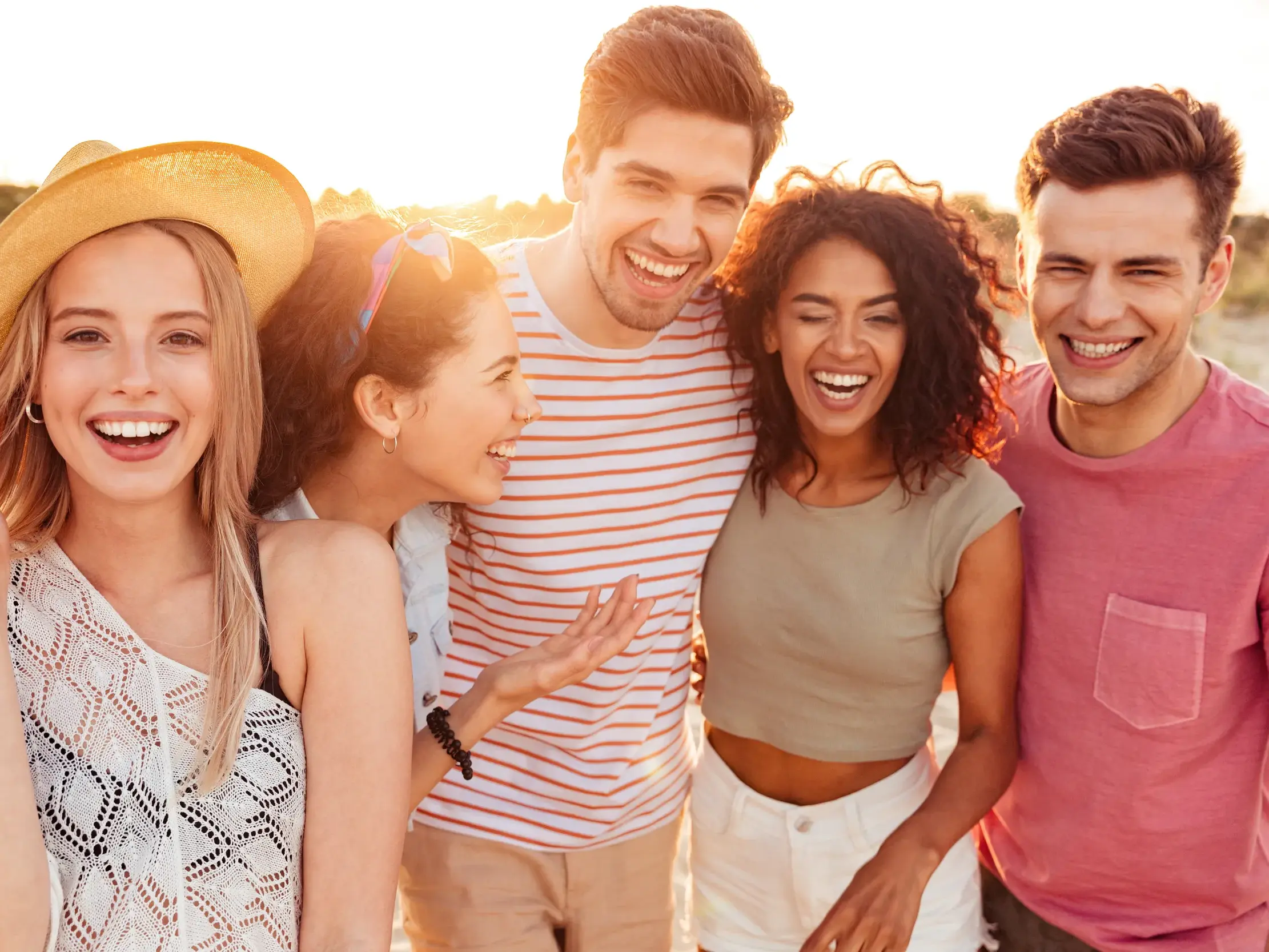 Fünf junge Freunde lachen ausgelassen am Strand bei Sonnenuntergang auf einer Gruppenreise für junge Leute in Griechenland.