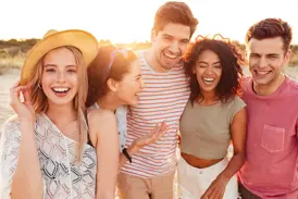 Fünf junge Freunde lachen ausgelassen am Strand bei Sonnenuntergang auf einer Gruppenreise für junge Leute in Griechenland.