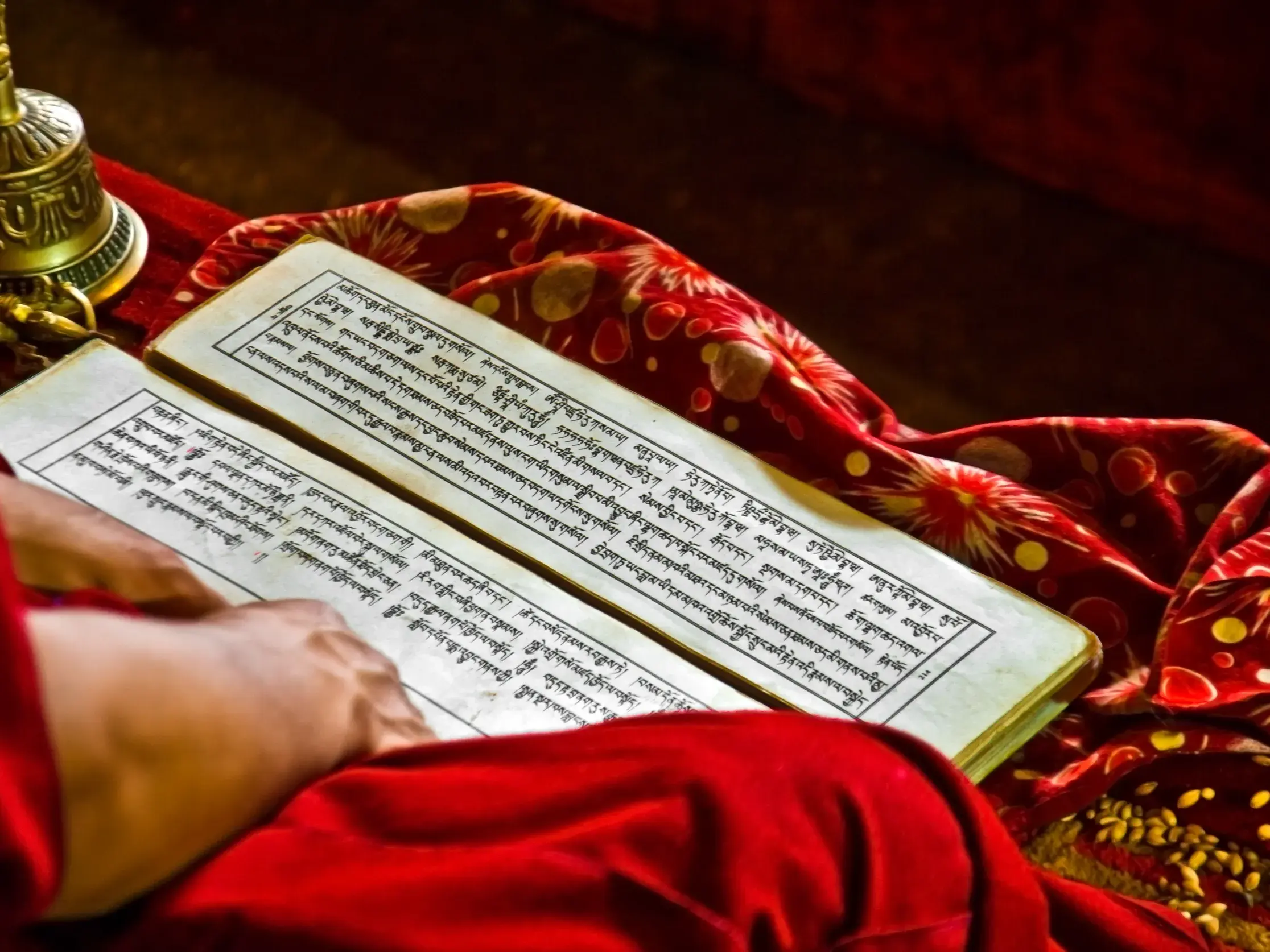 Ein tibetischer Mönch in roter Robe studiert bei warmem Licht eine heilige Schrift neben einer goldenen Ritualglocke in Tibet.