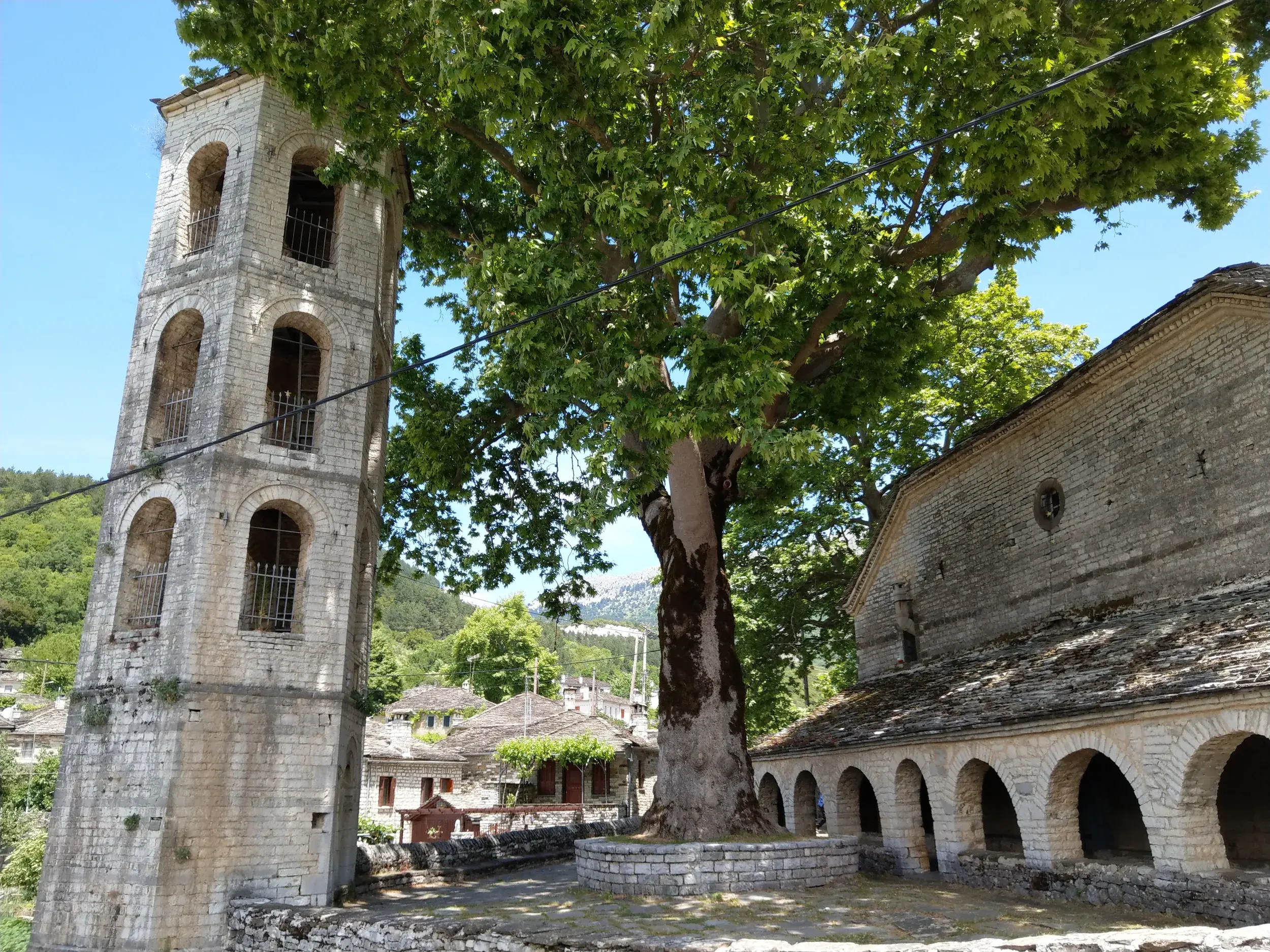 Ein hoher Glockenturm aus Stein neben einer großen Platane und einer Kirche im Bergdorf Megalo Papingo in der Zagori-Region.