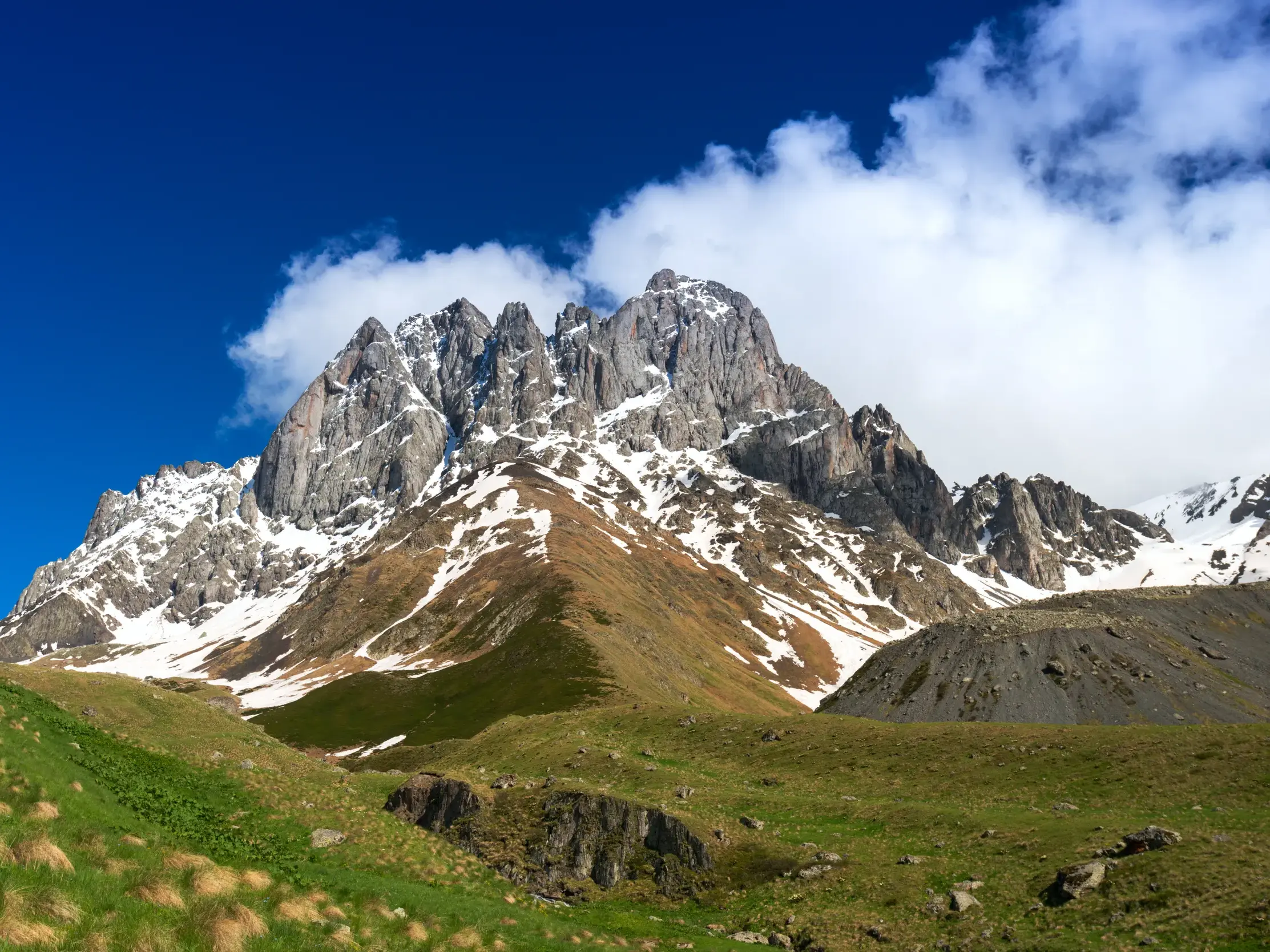 Grüne Hügel führen zum schneebedeckten, felsigen Chaukhi Massiv im Großen Kaukasus in Georgien unter strahlend blauem Himmel.