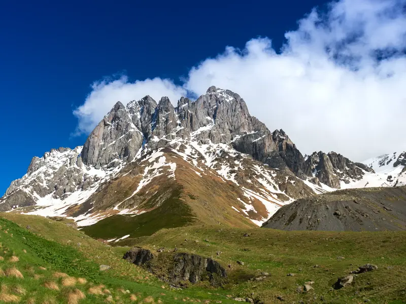 Grüne Hügel führen zum schneebedeckten, felsigen Chaukhi Massiv im Großen Kaukasus in Georgien unter strahlend blauem Himmel.