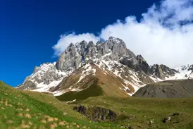 Grüne Hügel führen zum schneebedeckten, felsigen Chaukhi Massiv im Großen Kaukasus in Georgien unter strahlend blauem Himmel.