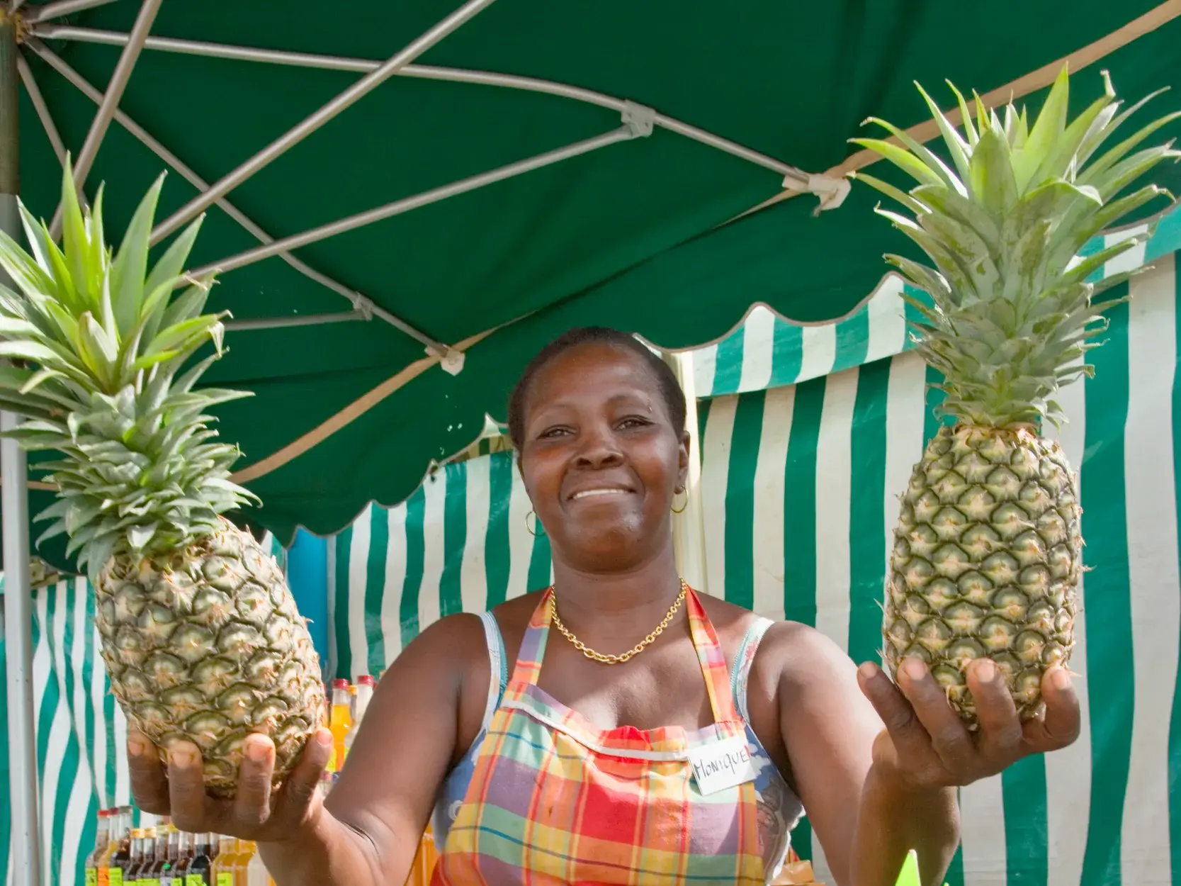 Eine freundliche Marktfrau in einer bunten Schürze präsentiert an ihrem Marktstand auf Martinique stolz zwei saftige Ananas.