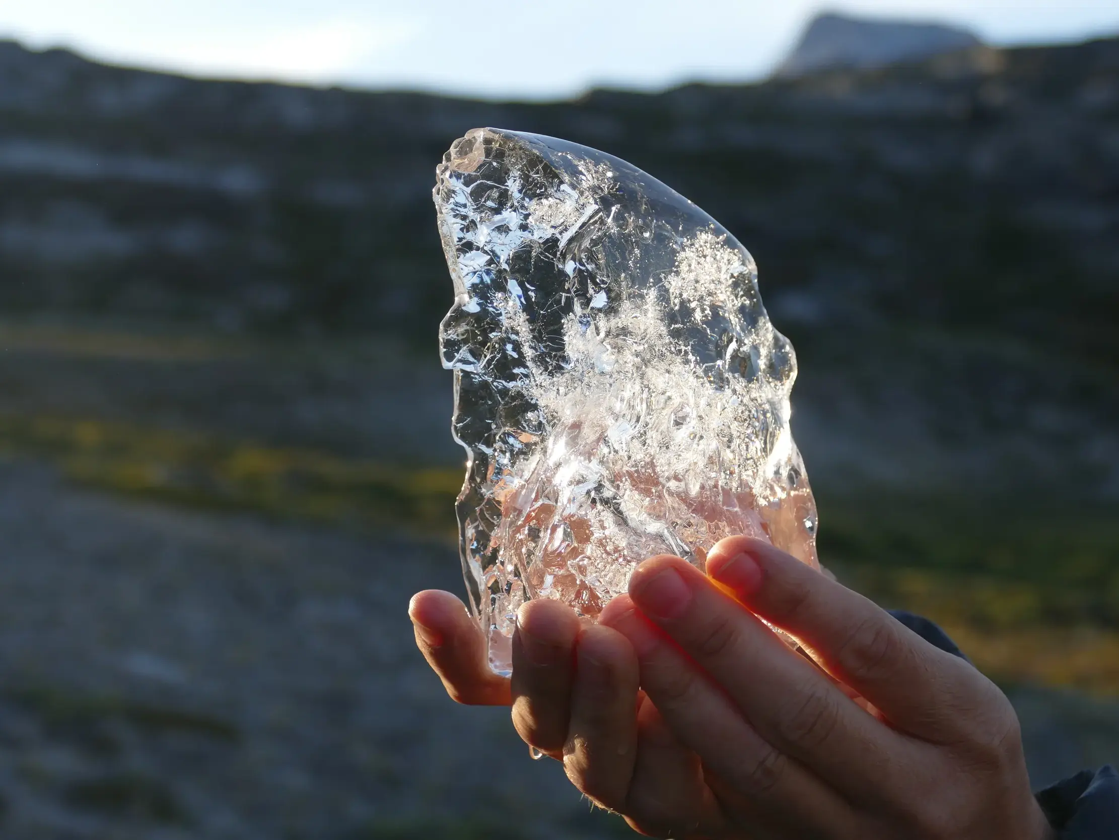 Hände halten ein Stück klares Gletschereis, das in der Sonne vor der unscharfen Felskulisse Grönlands funkelt.