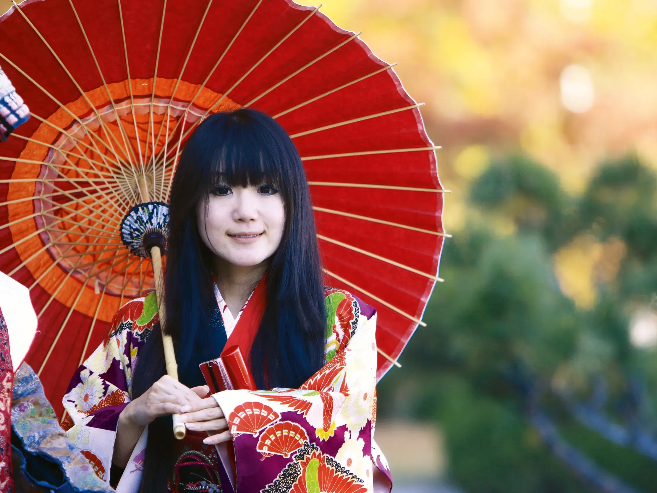 Junge Japanerin im bunten Kimono hält einen roten Papierschirm in einem sonnendurchfluteten Garten und lächelt.