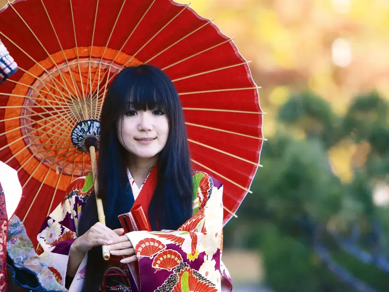 Junge Japanerin im bunten Kimono hält einen roten Papierschirm in einem sonnendurchfluteten Garten und lächelt.