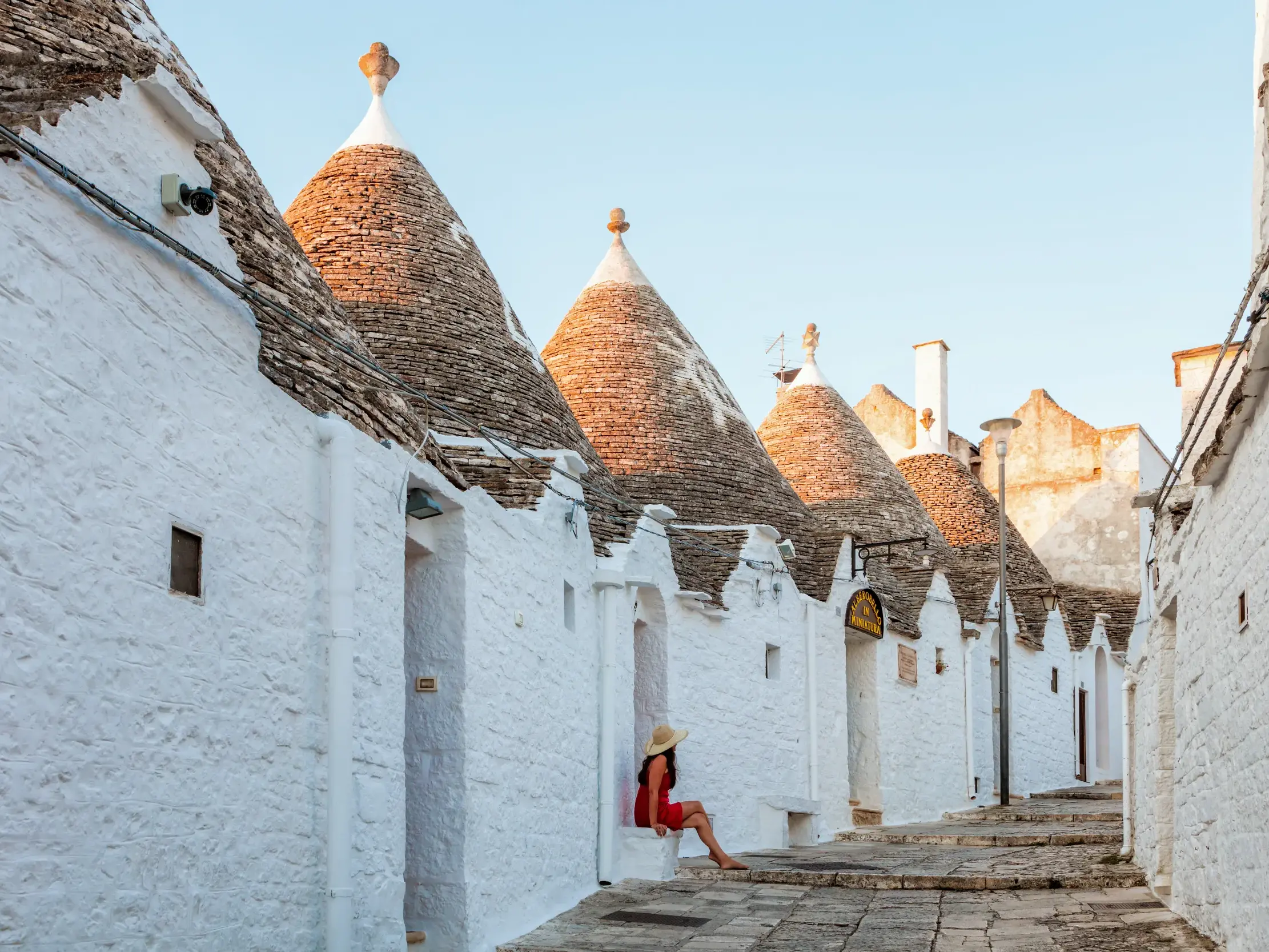 Frau in rotem Kleid sitzt in einer Gasse zwischen den charakteristischen Trulli-Rundhäusern von Alberobello in Apulien.