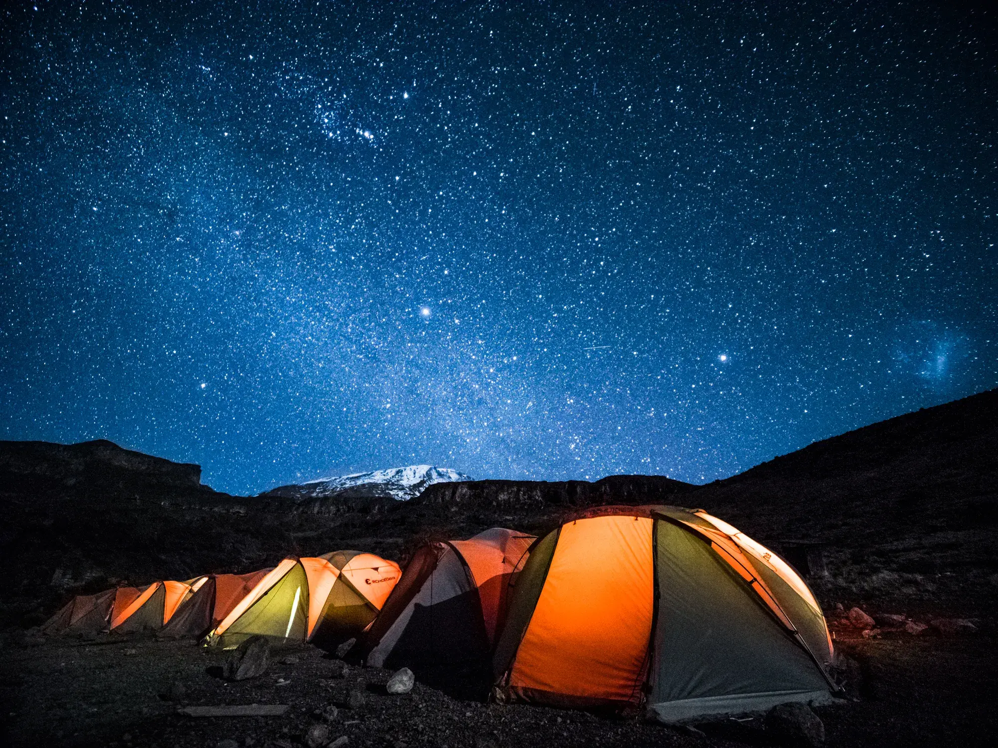 Eine Reihe leuchtender Zelte im Trekking-Camp am Kilimanjaro unter dem klaren Sternenhimmel mit Blick auf den Gipfel.