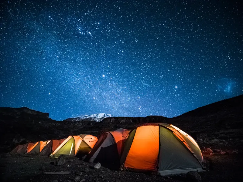 Eine Reihe leuchtender Zelte im Trekking-Camp am Kilimanjaro unter dem klaren Sternenhimmel mit Blick auf den Gipfel.