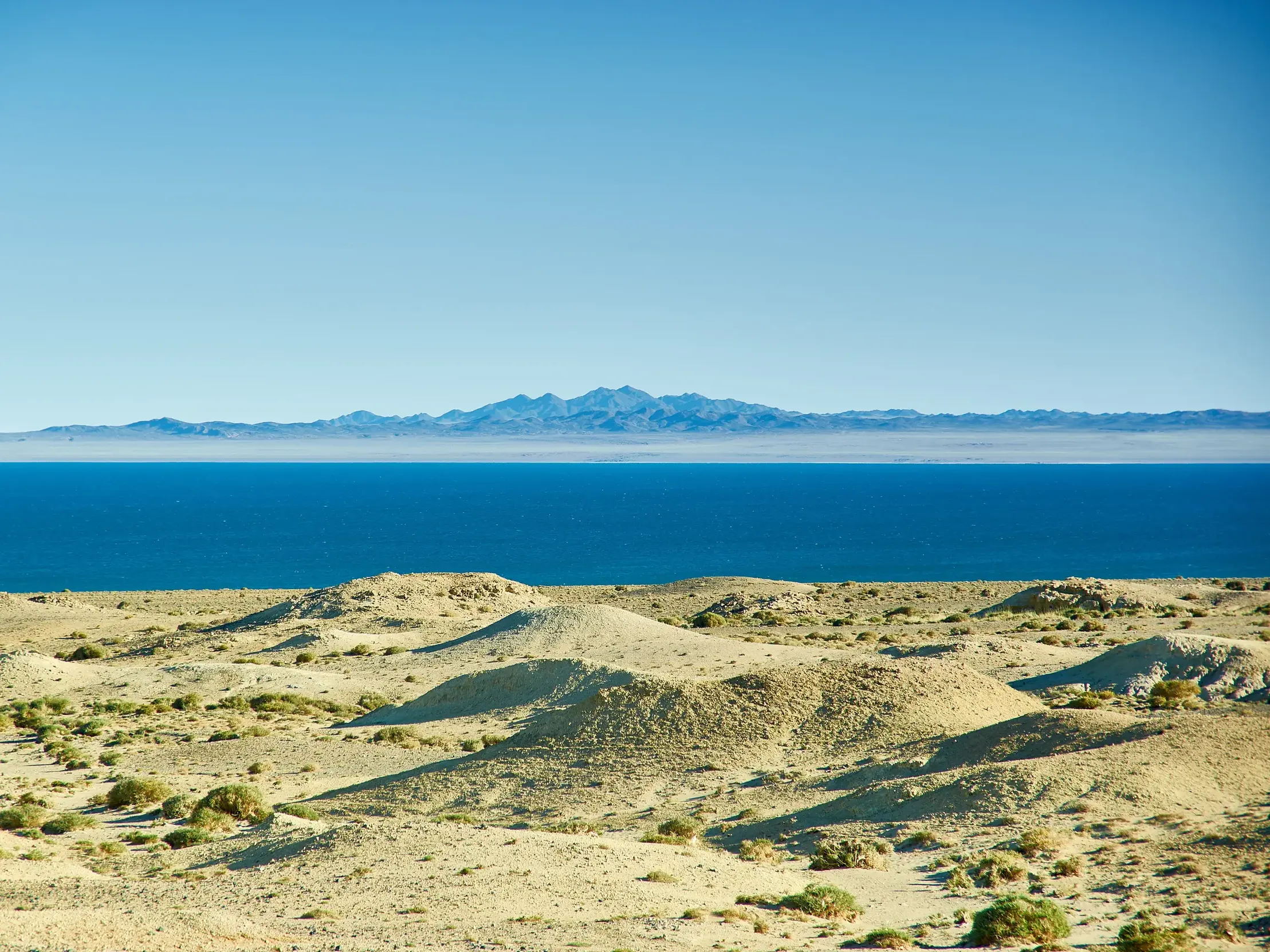 Sandige Hügel am Ufer des tiefblauen Uureg-Sees in der West-Mongolei, im Hintergrund eine ferne Gebirgskette.