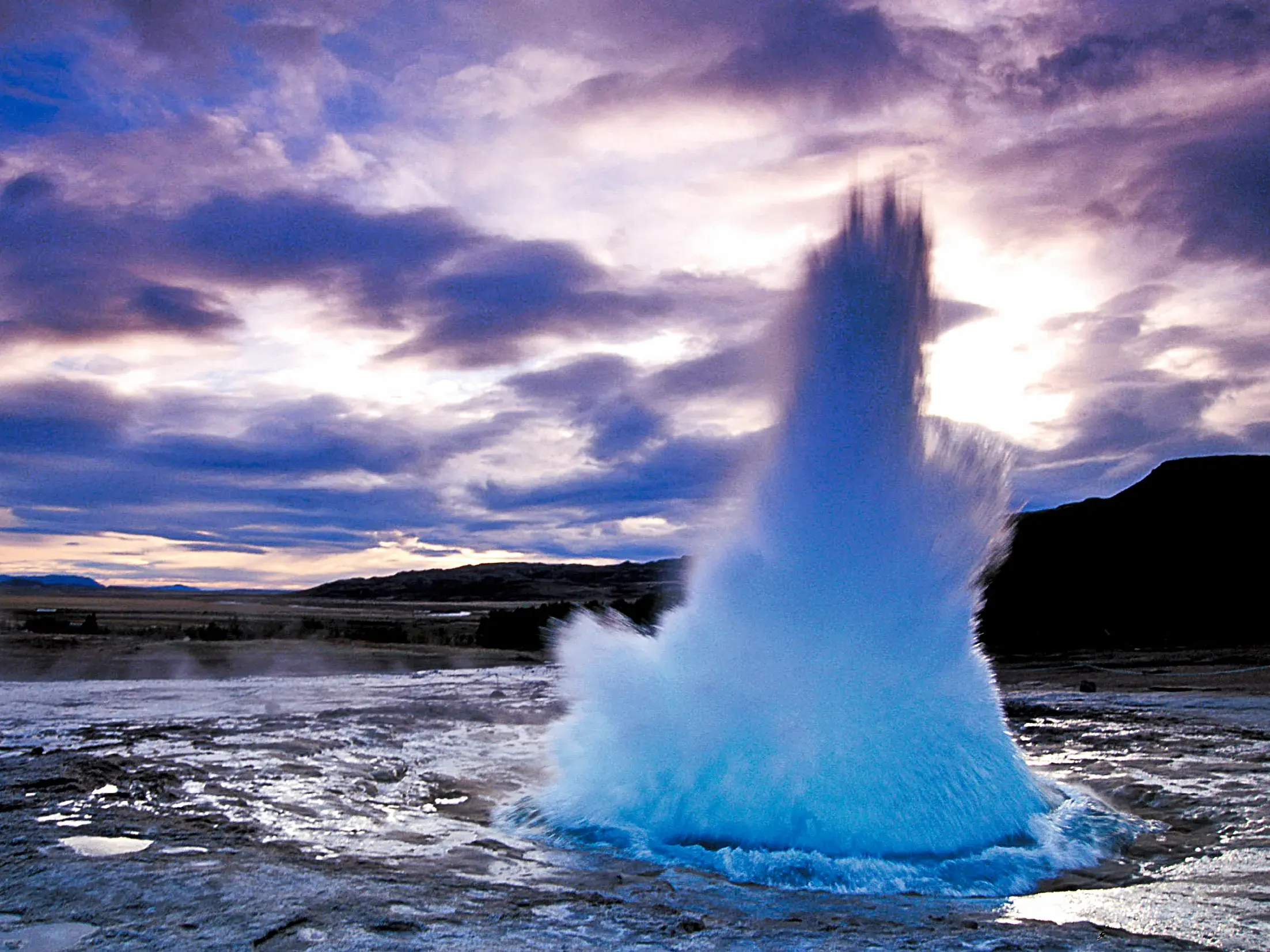 Der Geysir Strokkur auf Island bricht mit einer gewaltigen Wasserfontäne unter einem dramatischen, lila Wolkenhimmel aus.