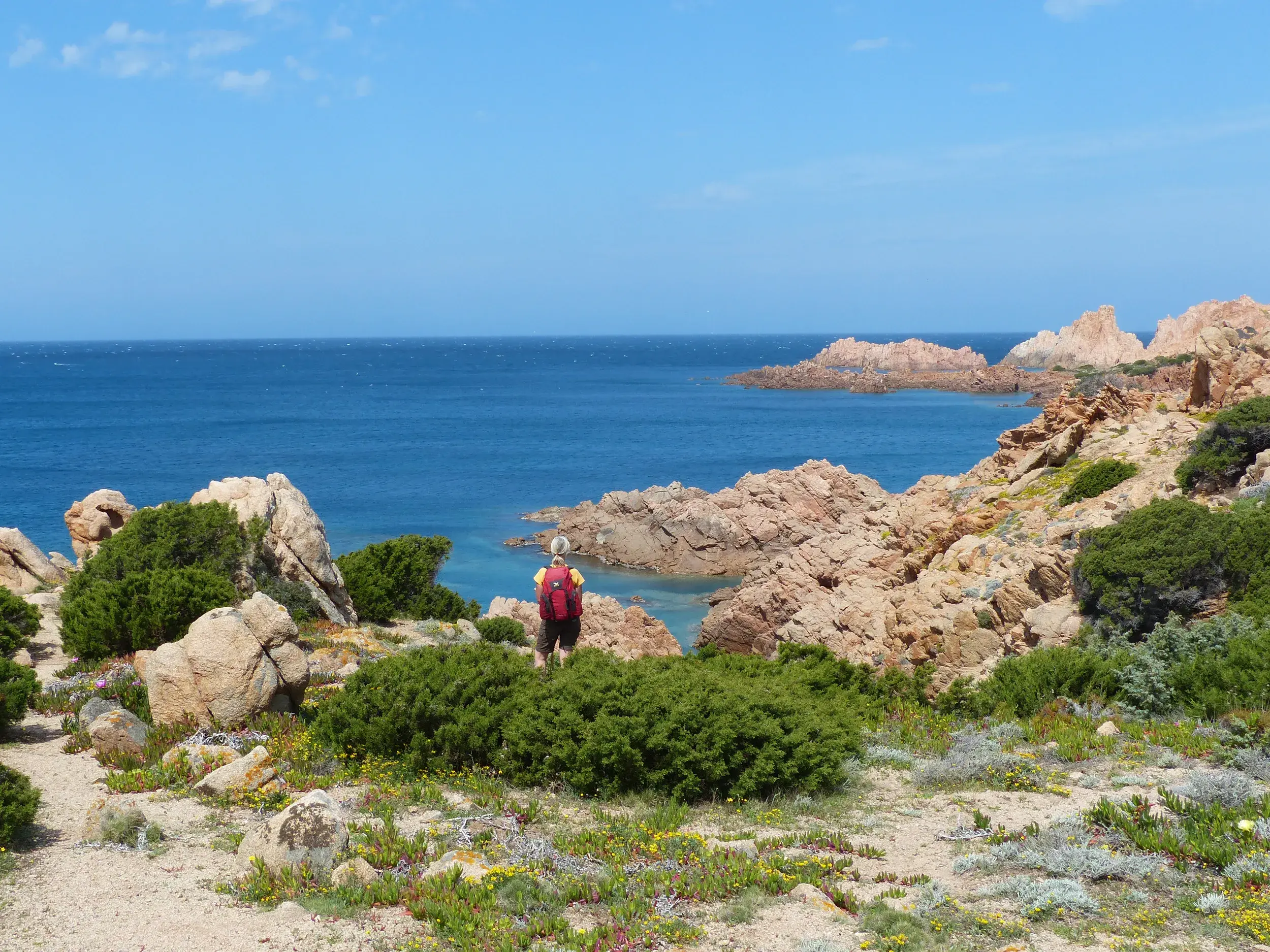Wanderer genießt auf einem Küstenpfad bei Isola Rossa auf Sardinien den Ausblick auf die leuchtend-roten Felsen und das Meer.