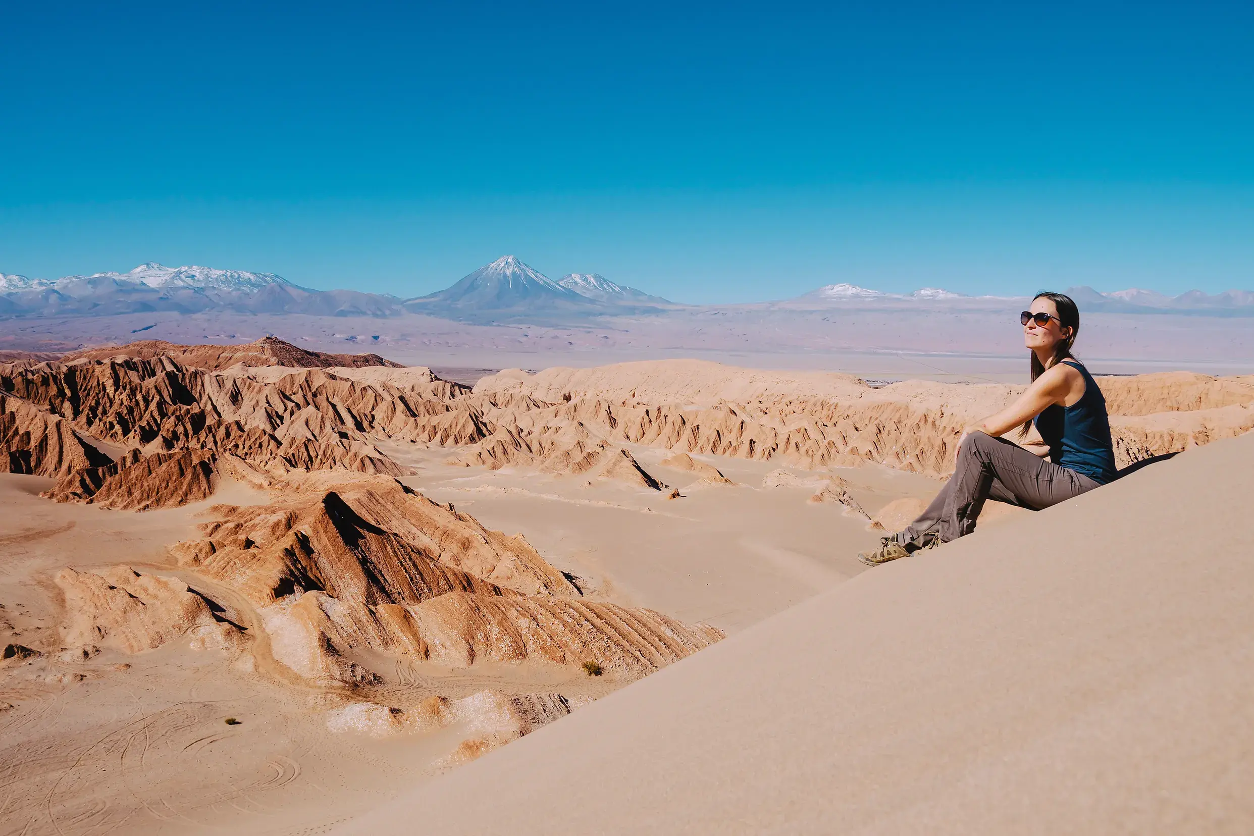 Eine junge Reisende genießt den Ausblick von einer Düne im Mondtal auf die Atacamawüste und Anden mit Vulkan Licancabur.