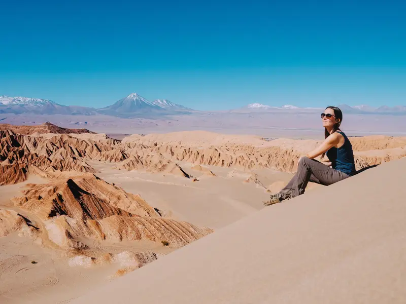 Eine junge Reisende genießt den Ausblick von einer Düne im Mondtal auf die Atacamawüste und Anden mit Vulkan Licancabur.