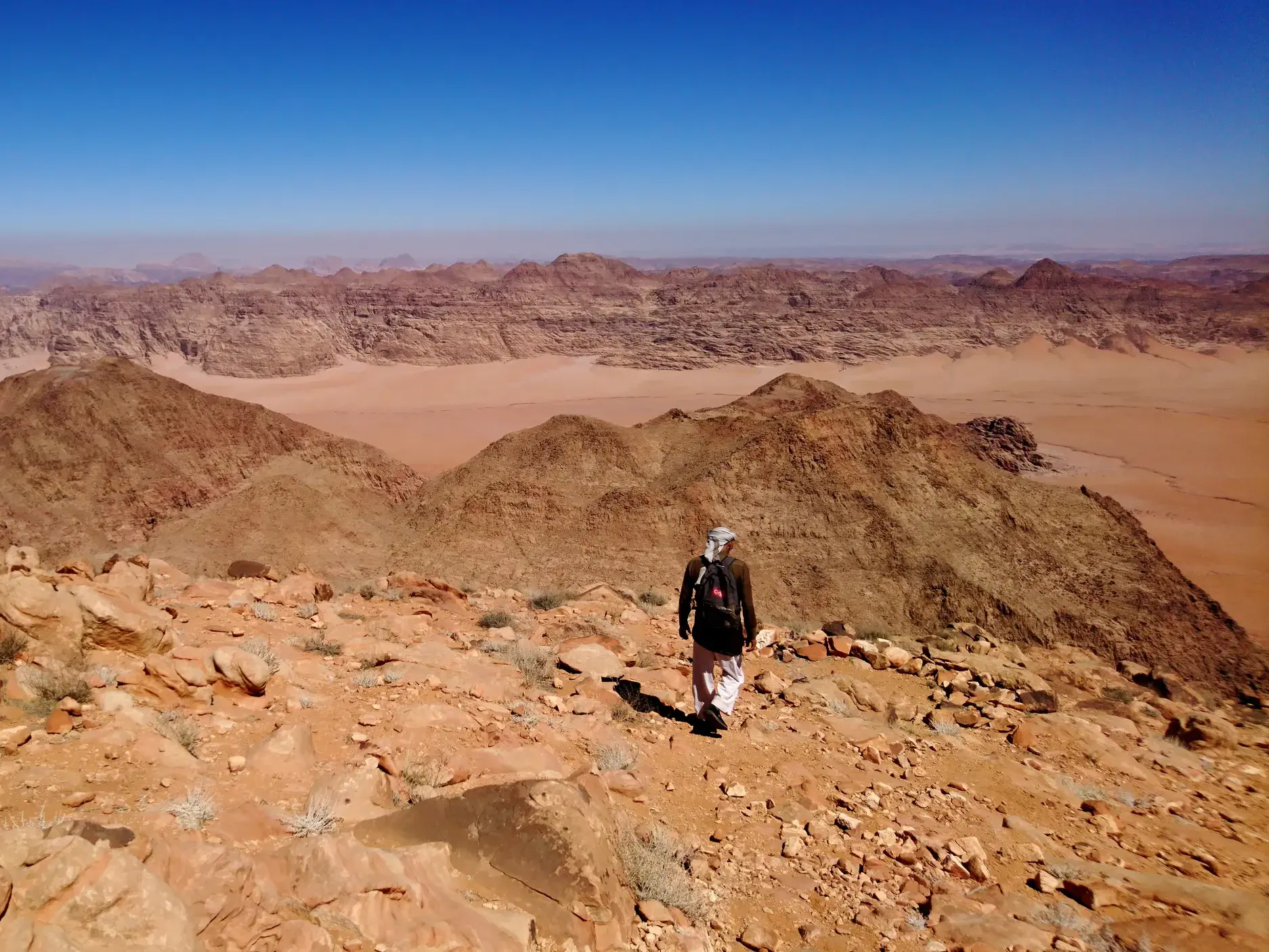 Ein Wanderer mit Rucksack beim Trekking auf dem Gipfel des Dschabal Umm ad-Dami mit weitem Blick über die felsige Wüstenlandschaft des Wadi Rum in Jordanien.
