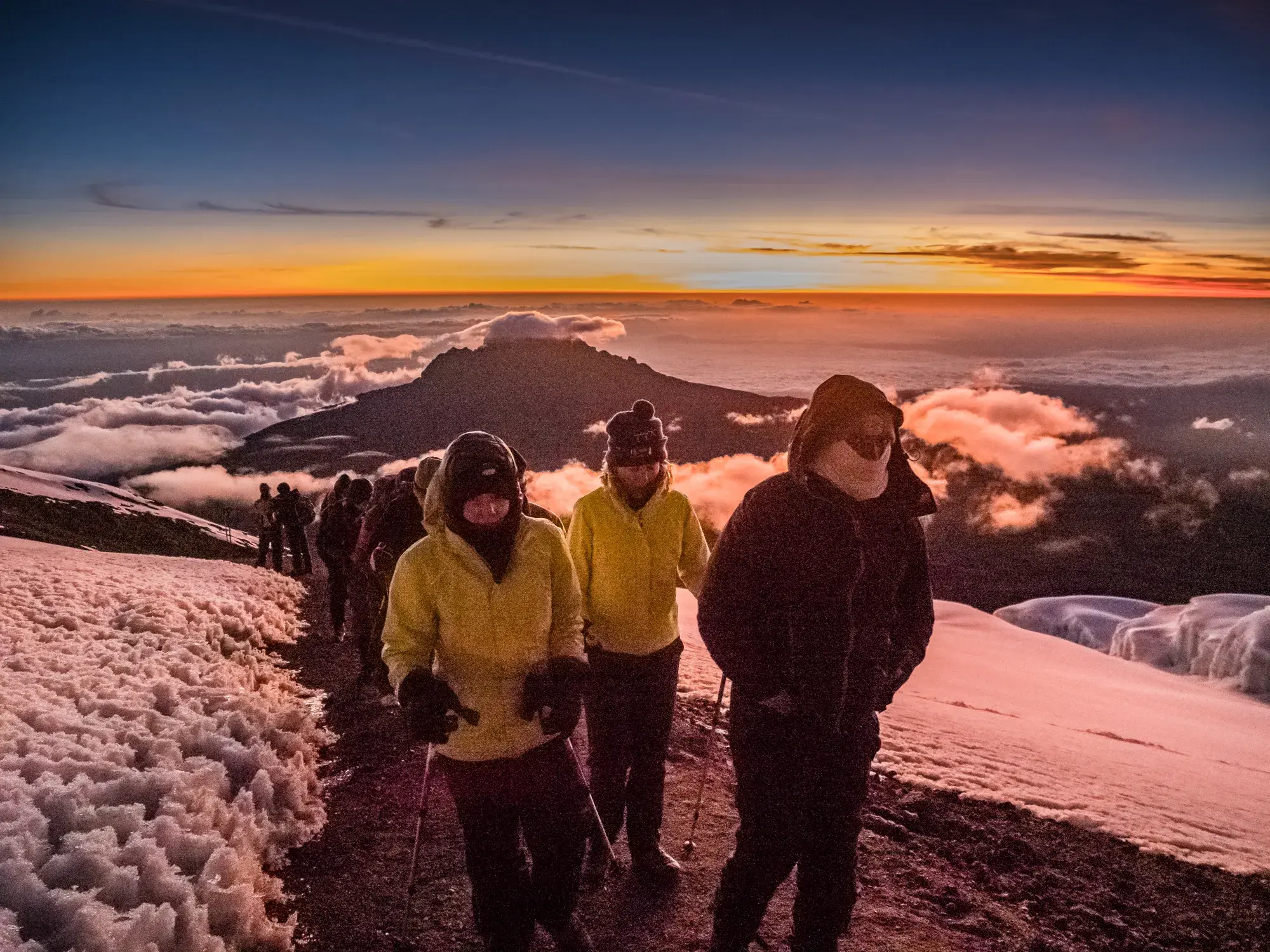 Eine Trekkinggruppe auf dem schneebedeckten Pfad am Kilimanjaro bei Sonnenaufgang mit Blick über die Wolken zum Mawenzi.
