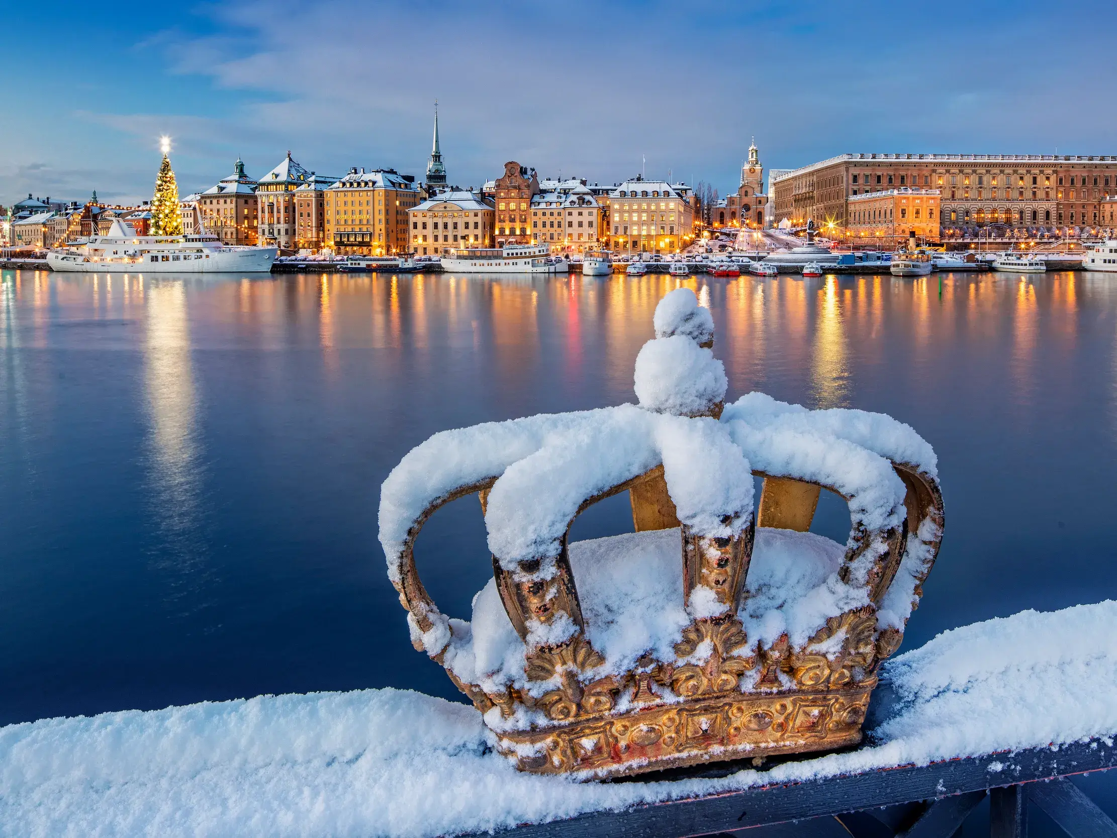 Eine schneebedeckte goldene Krone auf der Skeppsholmsbron-Brücke mit Blick auf die beleuchtete Altstadt Gamla Stan in Stockholm.
