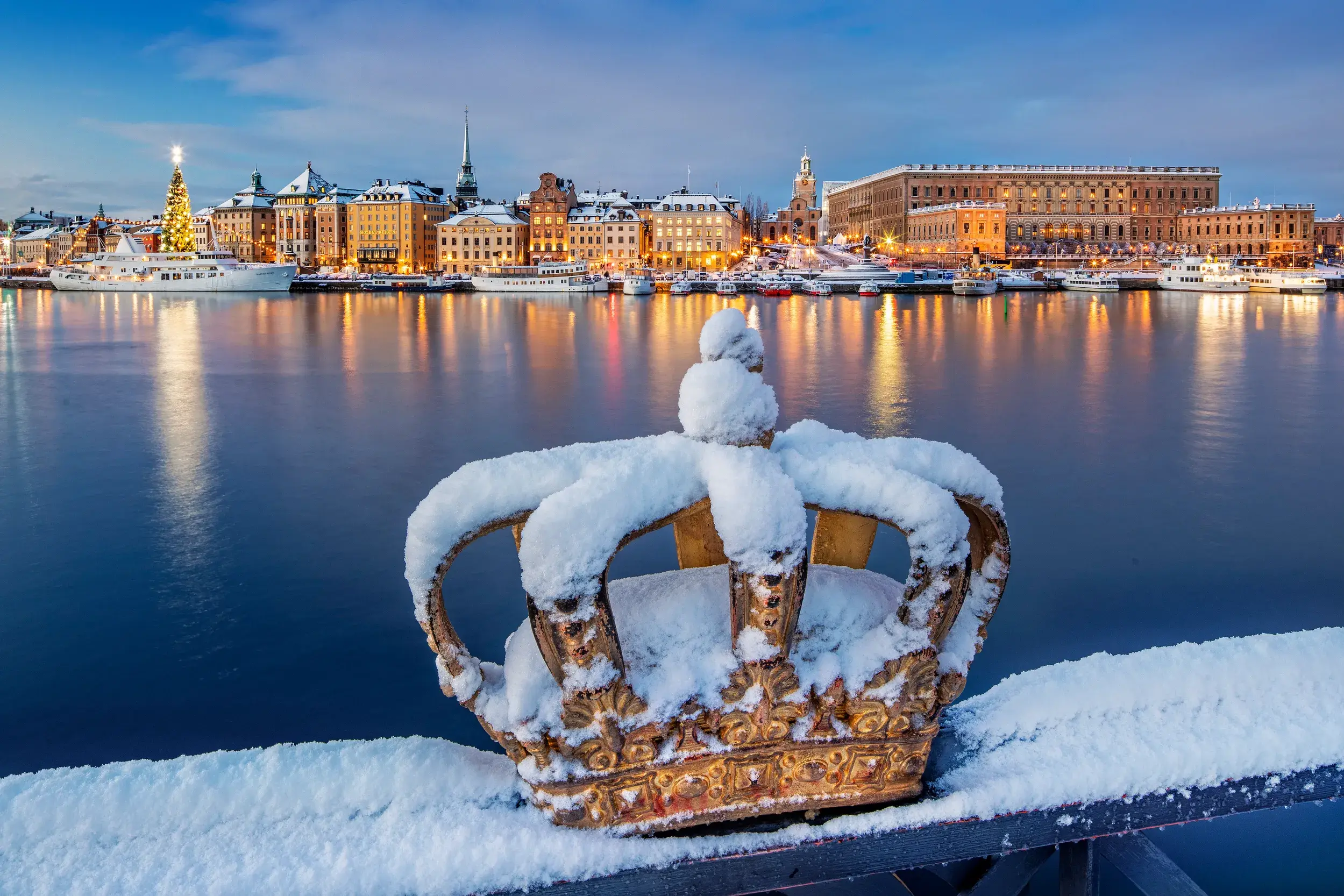 Eine schneebedeckte goldene Krone auf der Skeppsholmsbron-Brücke mit Blick auf die beleuchtete Altstadt Gamla Stan in Stockholm.