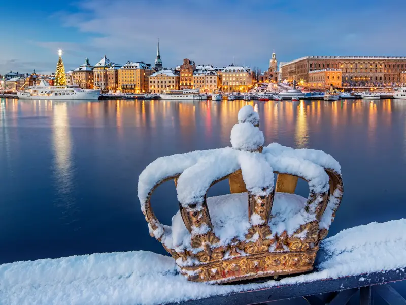 Eine schneebedeckte goldene Krone auf der Skeppsholmsbron-Brücke mit Blick auf die beleuchtete Altstadt Gamla Stan in Stockholm.