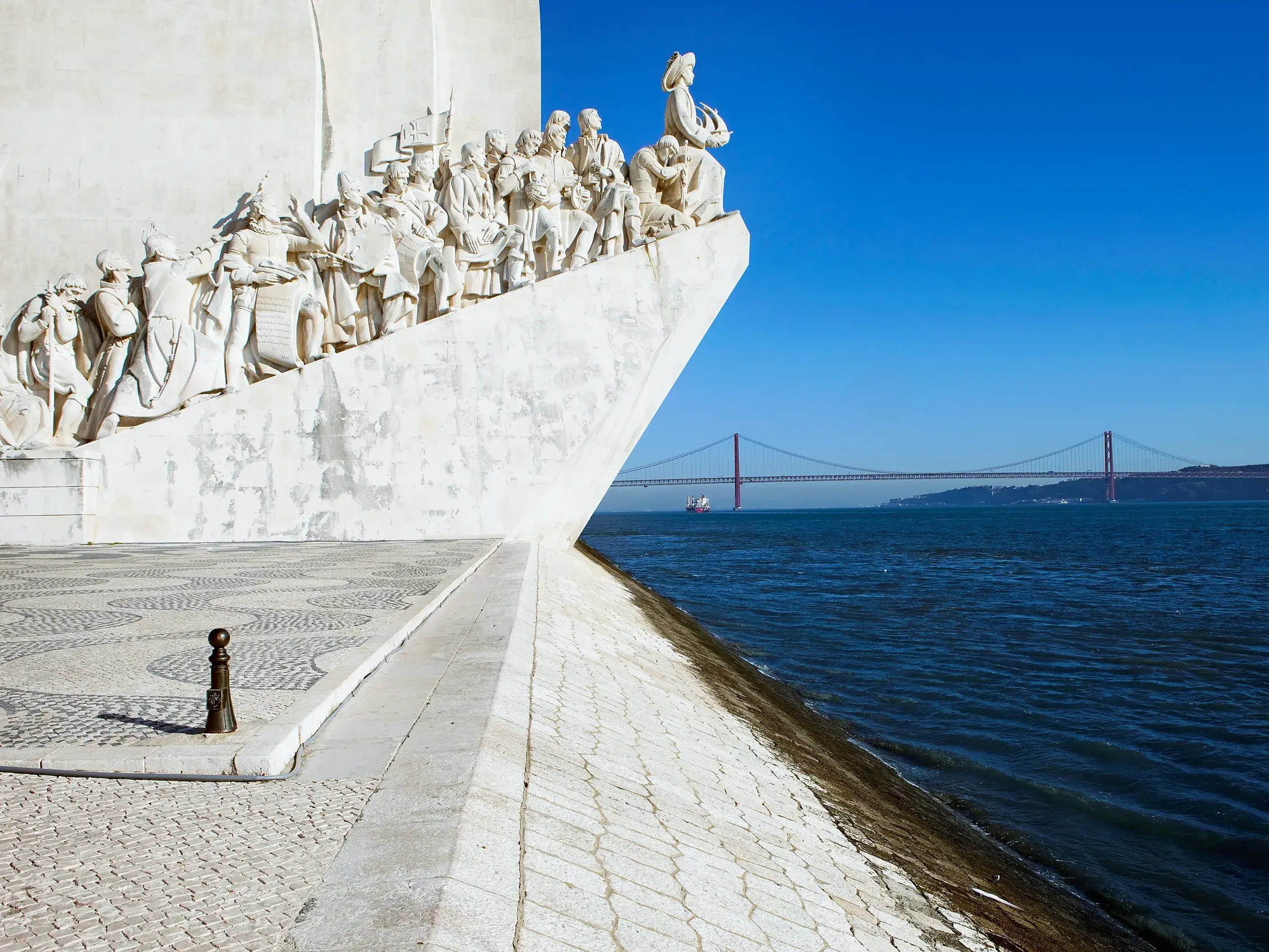 Das Seefahrer-Denkmal in Lissabon ragt am Ufer des Tejo in den blauen Himmel, im Hintergrund die rote Hängebrücke.