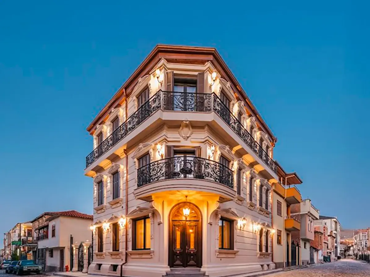 Beleuchtetes neoklassizistisches Gebäude des Artic Boutique Hotels in Korca mit schmiedeeisernen Balkonen vor blauem Abendhimmel.