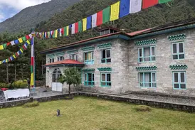 Außenansicht einer Komfort-Lodge der Mountain Lodges of Nepal in Phakding mit bunten Gebetsfahnen vor grünen Berghängen.