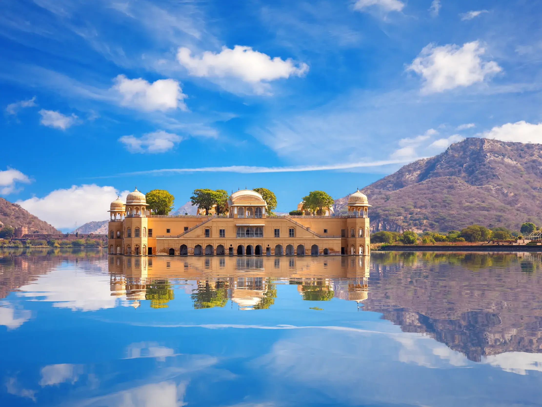 Der malerische Wasserpalast Jal Mahal in Jaipur spiegelt sich bei blauem Himmel im See vor den Aravalli-Hügeln.