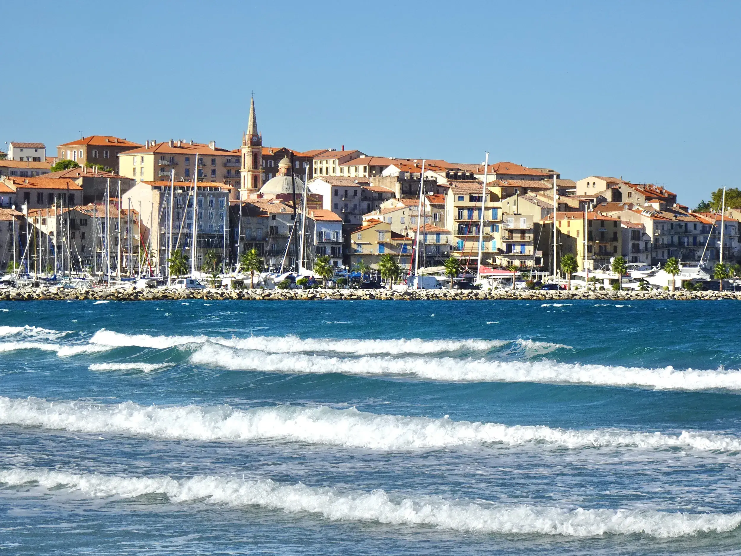 Sonnige Küstenansicht der Hafenstadt Calvi auf Korsika mit Wellengang im Meer und dem lebhaften Hafen voller Segelboote.