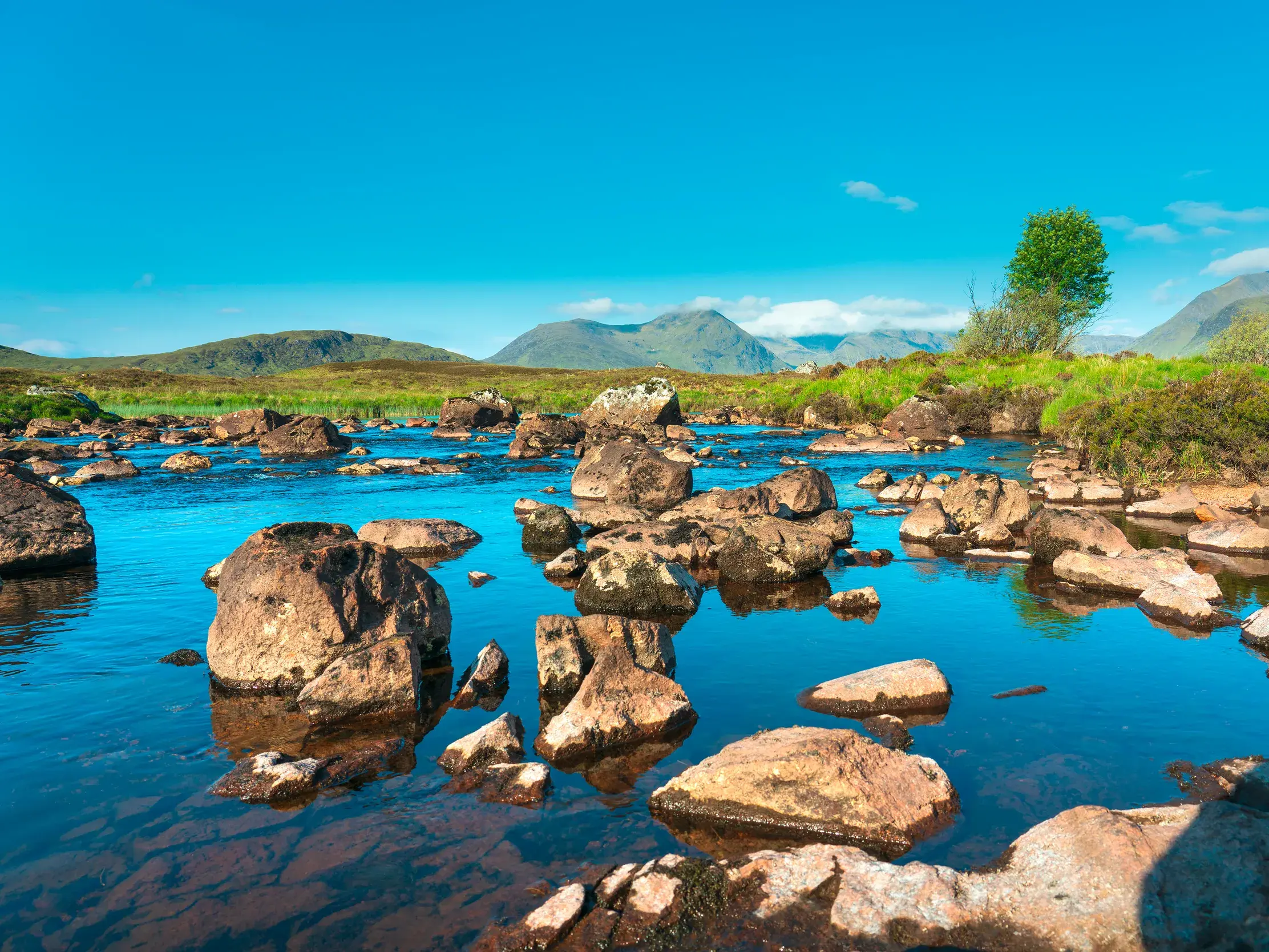 Ein klarer Fluss schlängelt sich durch die felsige Landschaft des Rannoch Moor in den schottischen Highlands unter blauem Himmel.