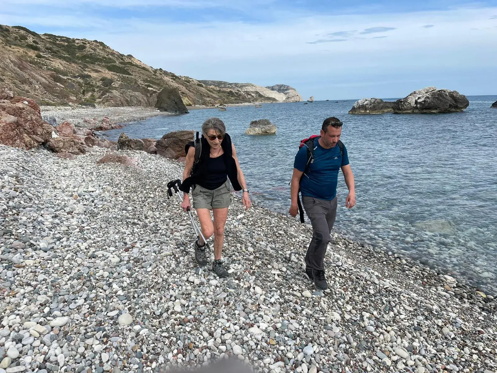 Eine Wanderin mit Stöcken und ein Mann mit Rucksack wandern auf einem Kieselstrand entlang der Felsküste Zyperns.