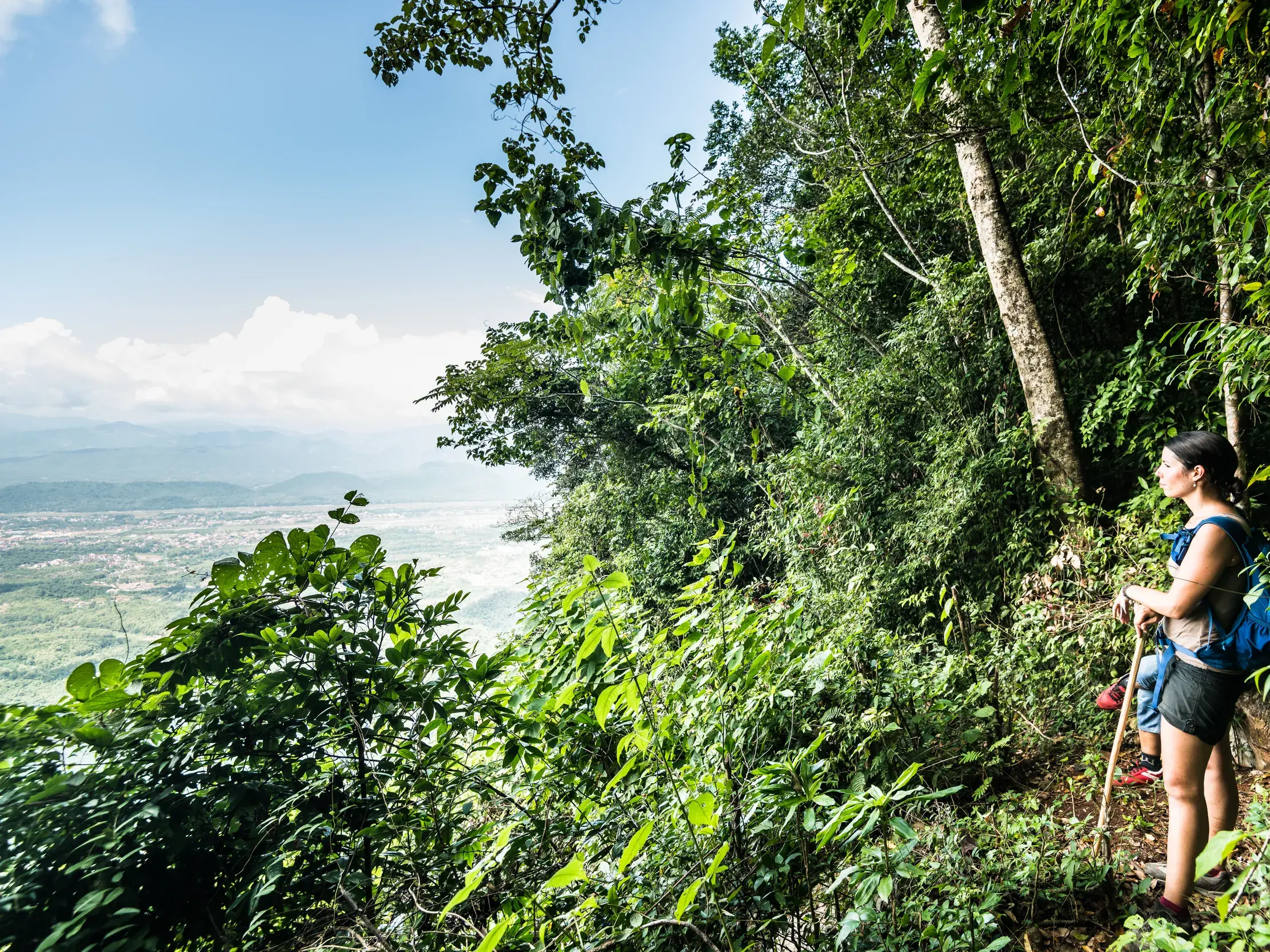 Eine Frau mit Rucksack und Wanderstock macht bei einer Trekking-Reise in Laos eine Pause auf einem schmalen, bewaldeten Pfad und blickt über das weite, grüne Tal.