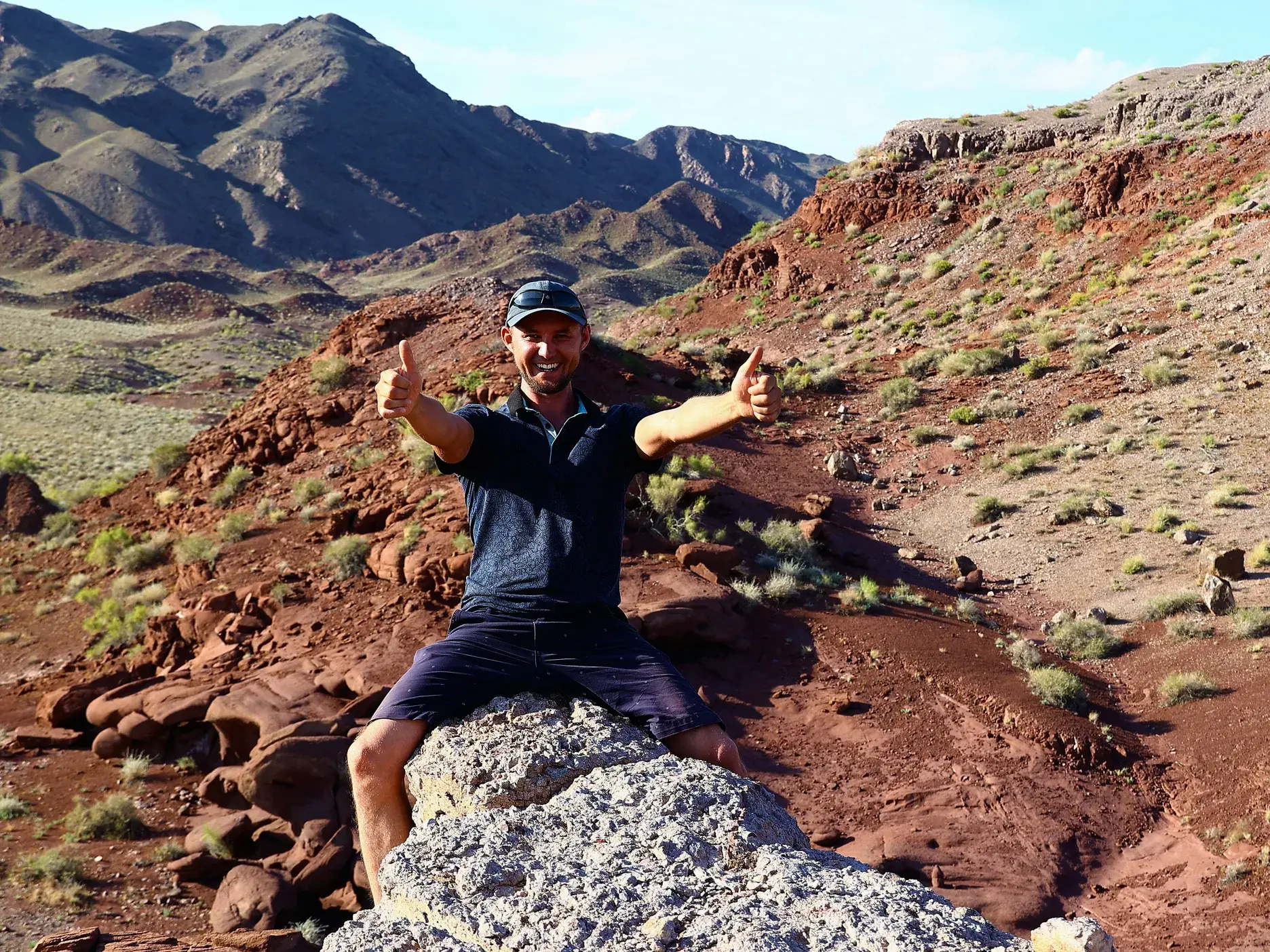 Ein Hauser-Reisender sitzt auf einem Felsen und gibt begeistert zwei Daumen hoch beim Trekking in den roten Aktau-Bergen Kasachstans.
