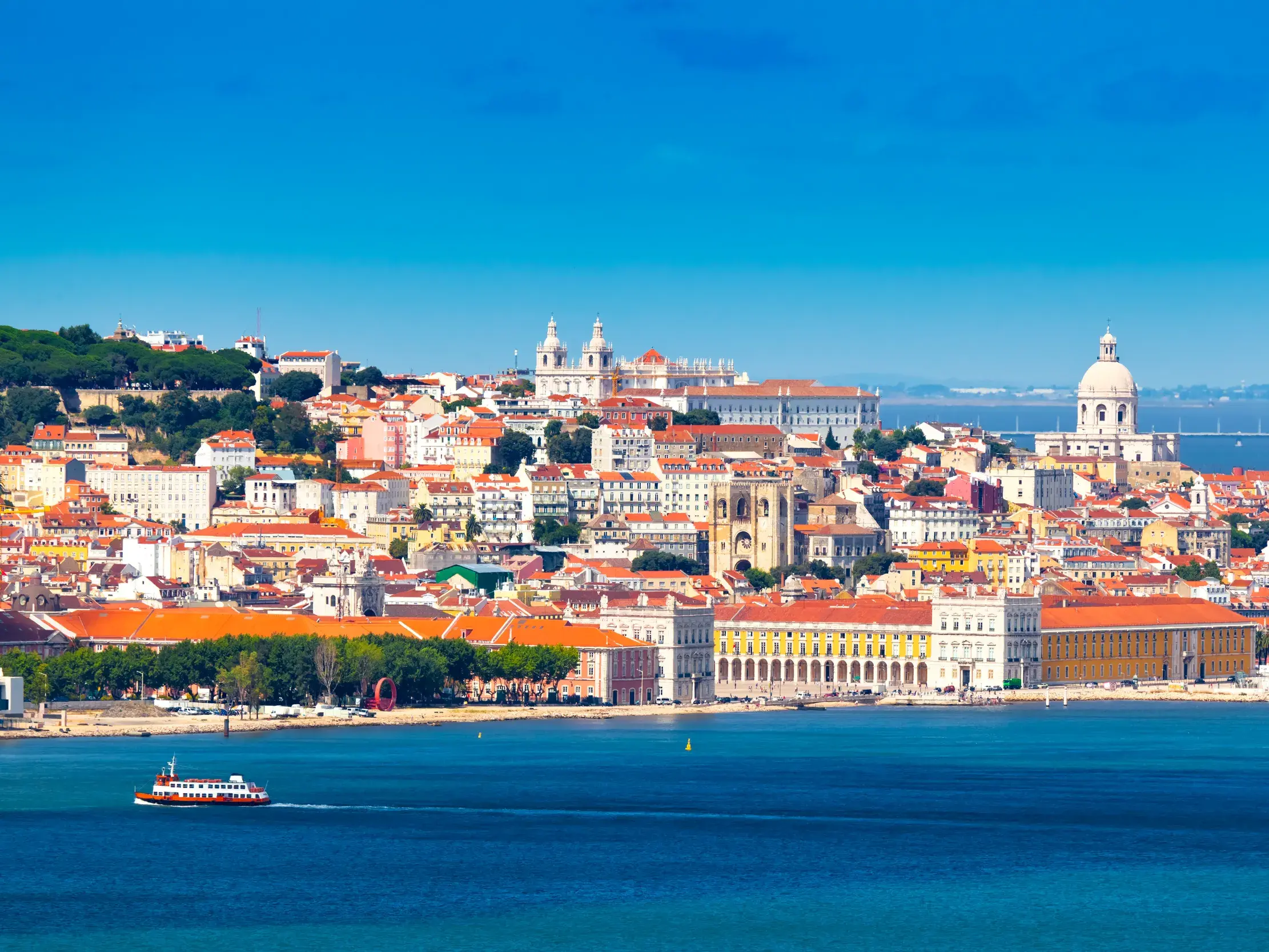 Malerischer Blick über den Rio Tejo auf die sonnige Altstadt Alfama in Lissabon mit einer Fähre im Vordergrund.