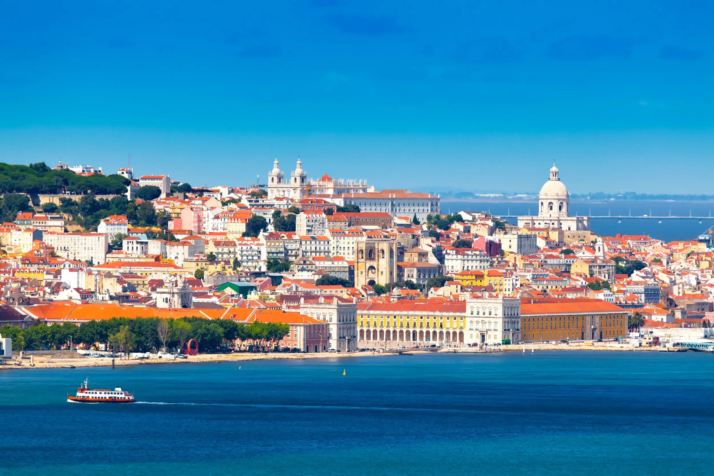 Malerischer Blick über den Rio Tejo auf die sonnige Altstadt Alfama in Lissabon mit einer Fähre im Vordergrund.